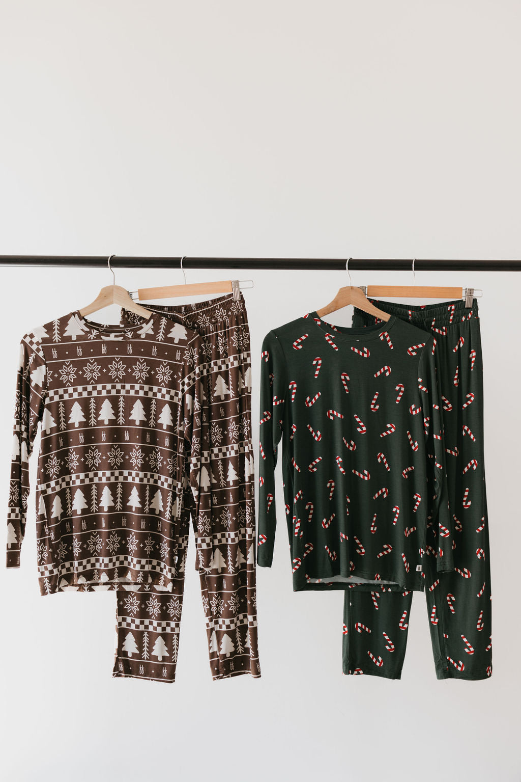 On a rack hang two sets of holiday-themed pajamas. The set on the left is crafted from bamboo fabric, featuring brown material with white festive patterns of snowflakes and trees. The set on the right showcases Forever French Baby's "Men's Bamboo Pajamas | Forever Fair Isle" in dark green, adorned with red and white candy cane patterns, providing a cozy hypo-allergenic choice for the season.