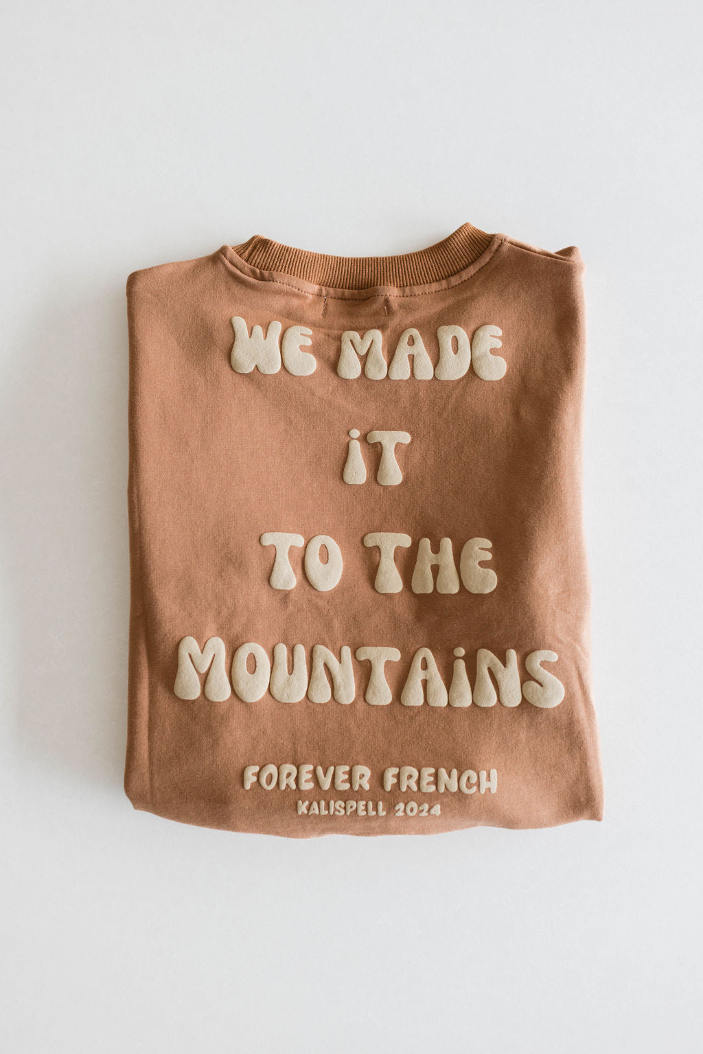 A folded brown sweatshirt from Forever French Baby, titled "Children's Sweatshirt | Made It to The Mountains," features the text "We Made It To The Mountains" in large, cream-colored letters. Below that, it reads "Forever French Kalispell 2024" in smaller cream-colored letters. Crafted from 95% Cotton and Elastane for a perfect fit, this cozy piece is available for local pickup.