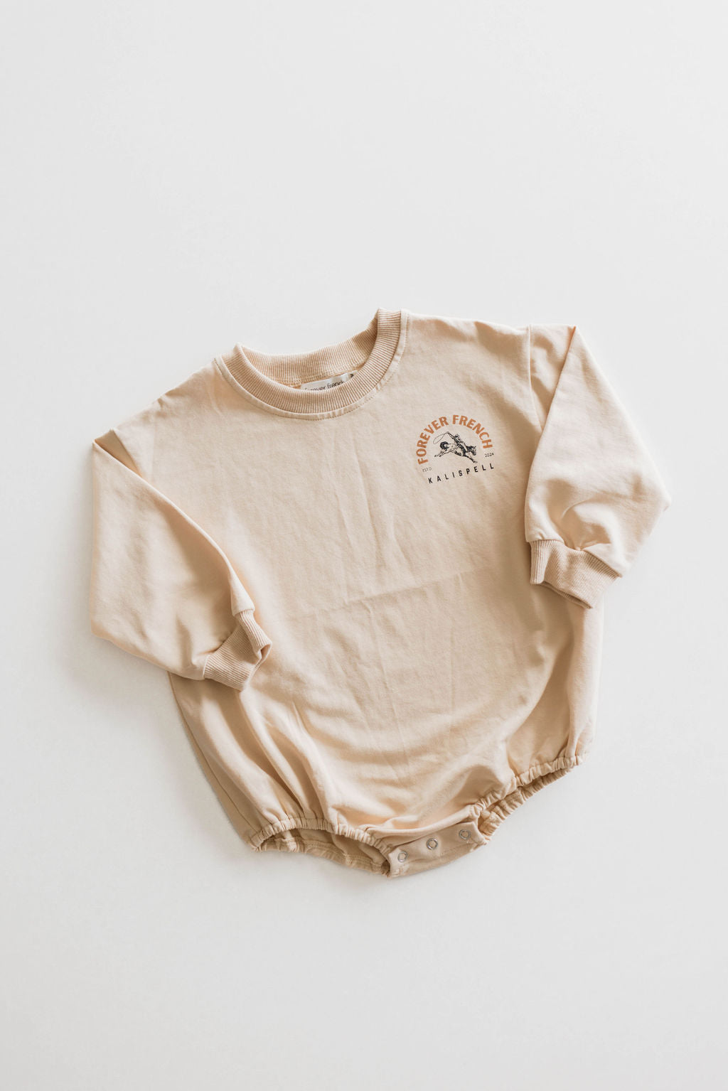 A beige long-sleeved baby romper from Forever French Baby, named Kalispell, is crafted from 95% cotton and elastane. It features the text "Forever Freewheel California" along with a small graphic of the sun on the chest. Convenient snap closures at the bottom ensure ease of use. Please note that this item is not available for local pickup.