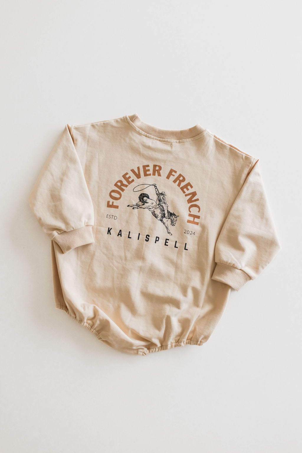 Introducing the Baby Romper | Kalispell by forever French baby: a beige long-sleeve onesie crafted from 95% cotton and elastane. This adorable piece features the printed text "FOREVER FRENCH" above an illustration of a man riding a bicycle, designed in brown and light orange hues. It also includes "KALISPELL" with "ESTD 2024" beneath it. Please note, this item is not available for local pickup.