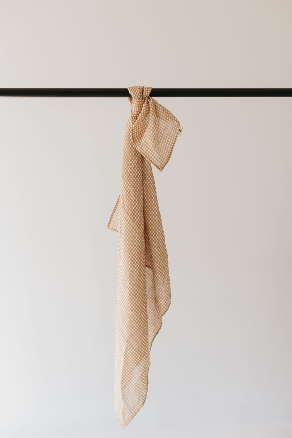 A beige and white Muslin Swaddle in the Golden Gingham pattern by forever french baby is draped over a black rod against a plain light background. The fabric hangs loosely, highlighting its soft texture reminiscent of a cozy scarf.