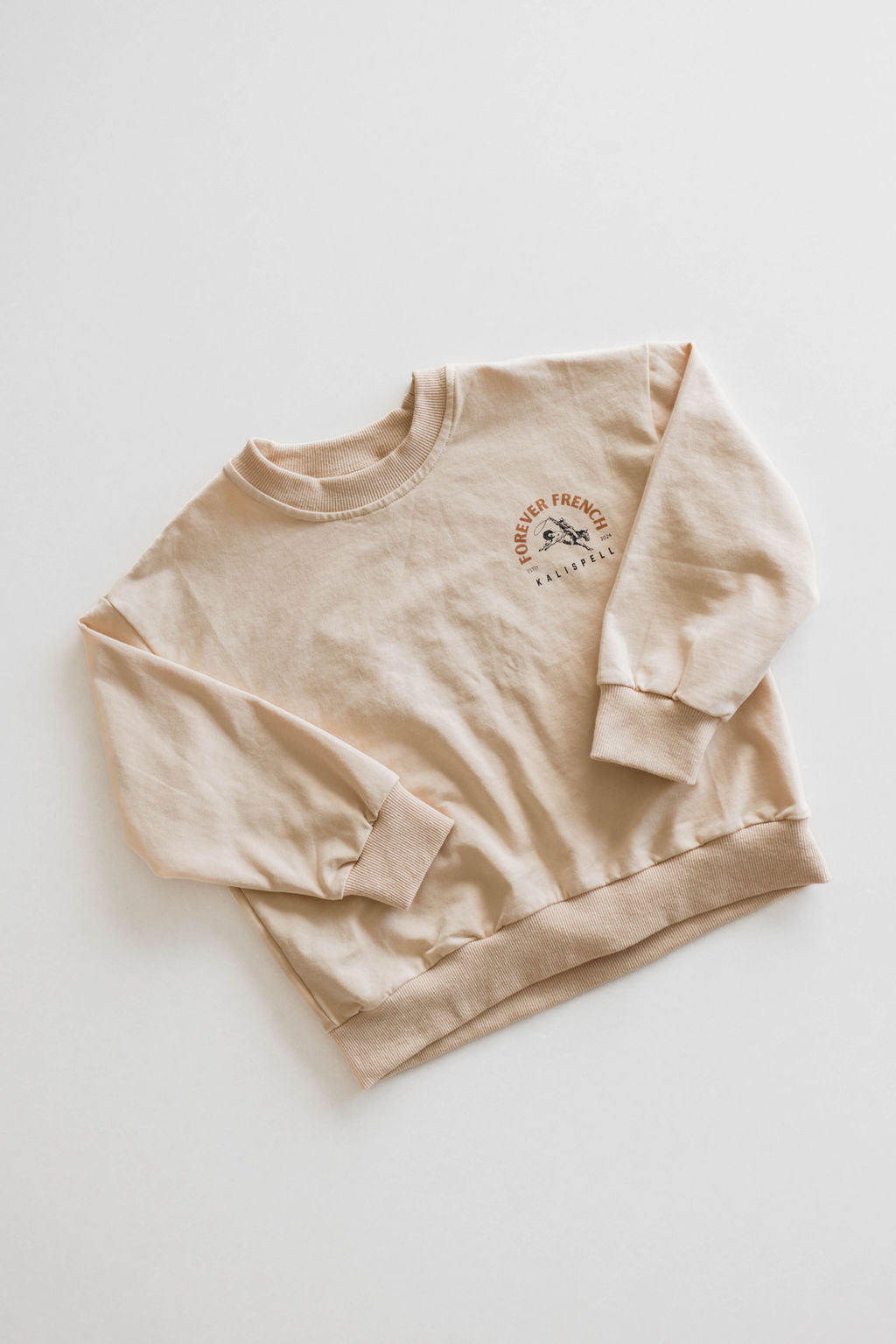 The Children's Sweatshirt by forever French baby is displayed flat on a white surface. This beige Kalispell sweatshirt features long sleeves, a ribbed waistband and cuffs, and a small printed design on the chest with text that reads "Forever Friends." Made from 95% cotton with elastane for added stretch, it is available for local pickup.