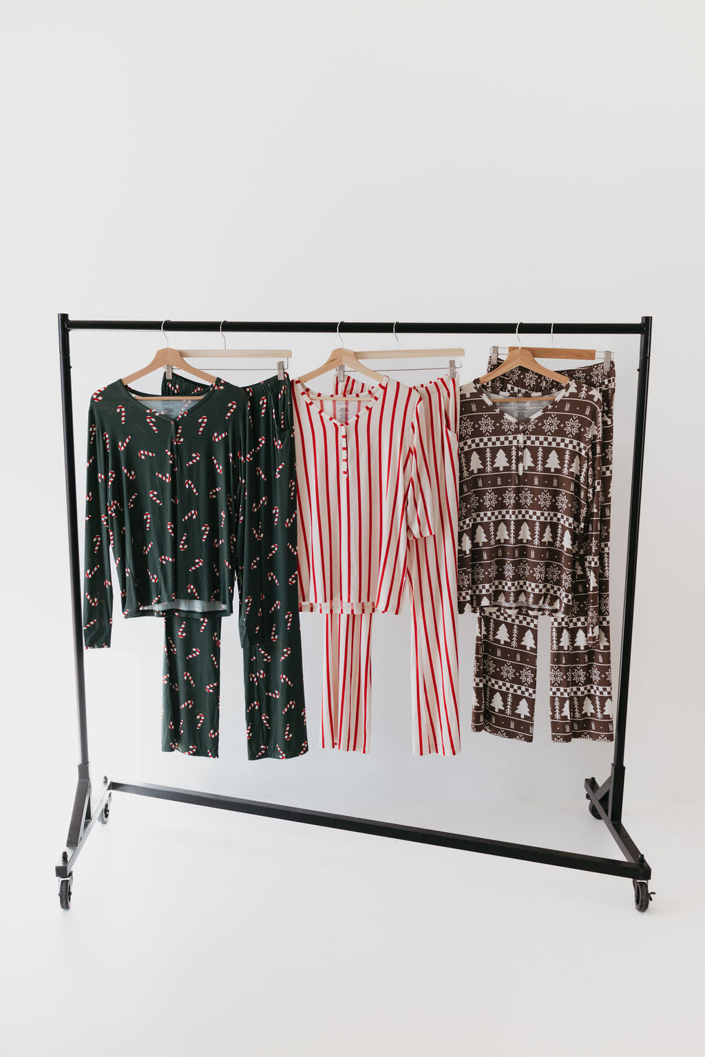 A clothing rack holds three pairs of pajamas, featuring a set of Women's Bamboo Pajamas from the "Forever Fair Isle" collection by forever french baby. The first pair is dark green with candy cane patterns, the second pair is red and white striped, and the third exhibits a brown and white winter design. All are displayed against a simple, plain white background.