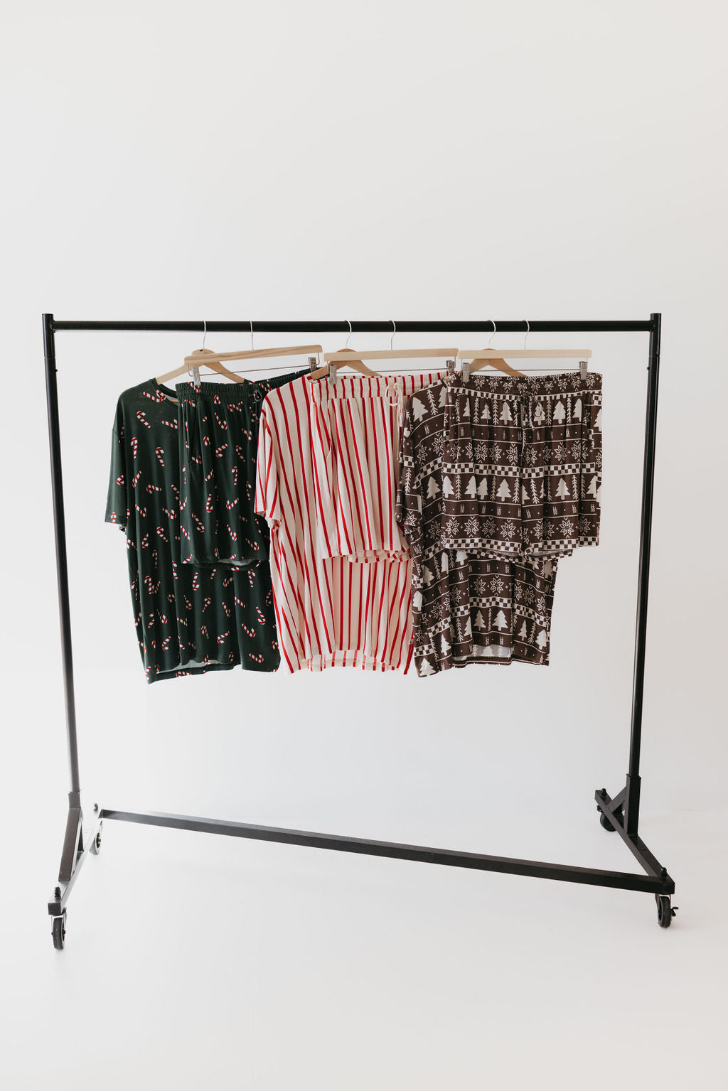 On a black clothing rack, three garments are displayed: the Candy Cane Lane Short Sleeve Men's Bamboo Pajamas by forever french baby in a black and red print, one with elegant white and red stripes, and another featuring a brown and white pattern. These pieces are made from breathable bamboo fabric for exceptional comfort against the plain white background.