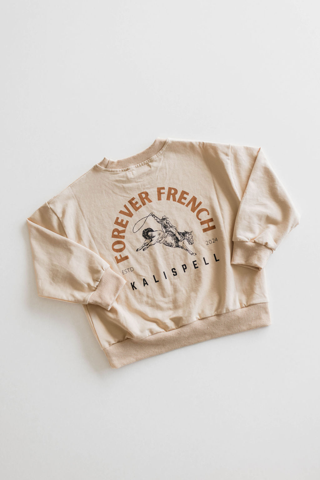 The Children's Sweatshirt by forever French baby, made of 95% cotton, is in beige and features the bold text "FOREVER FRENCH" along with "KALISPELL" and numbers "1870 2024" in smaller print. The design includes an illustration of a person on horseback. The sweatshirt is laid flat on a white background and is available for local pickup.