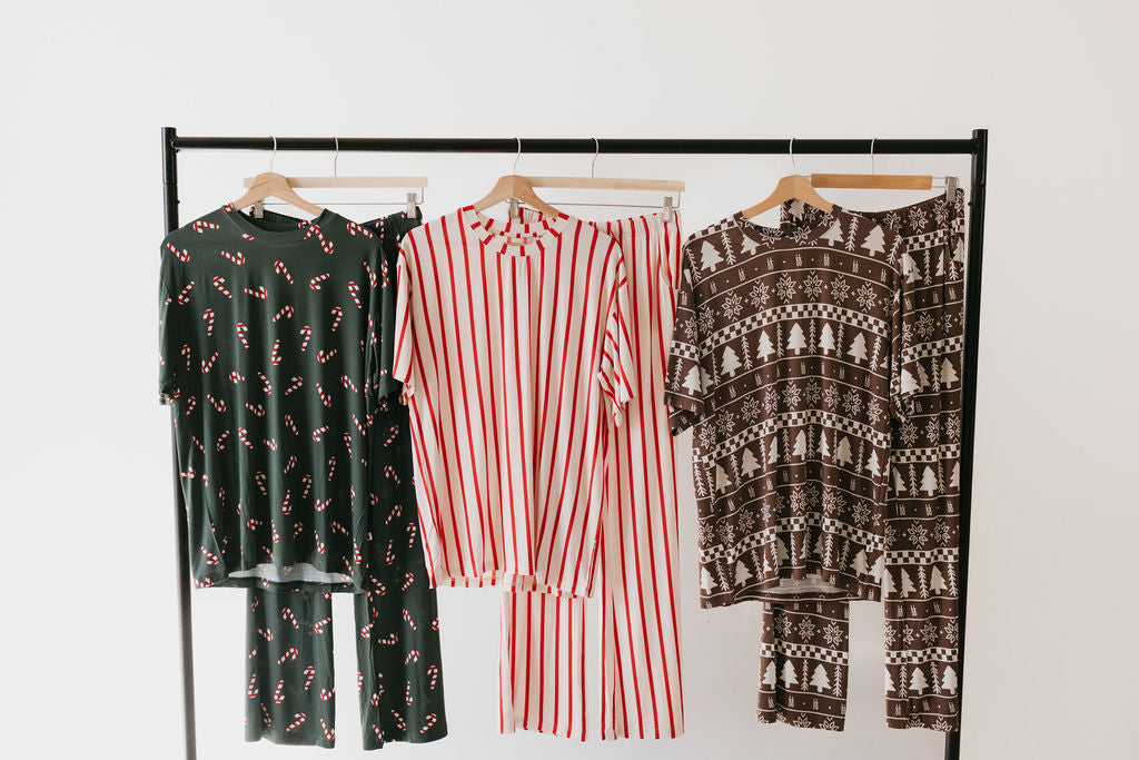 The black clothing rack holds four Short Sleeve Women's Bamboo Pajama sets from forever french baby. These cozy, oversized designs exhibit a variety of festive patterns, including candy canes on green fabric, red and white stripes, and brown adorned with Christmas trees and snowflakes, offering comfort with a holiday flair.