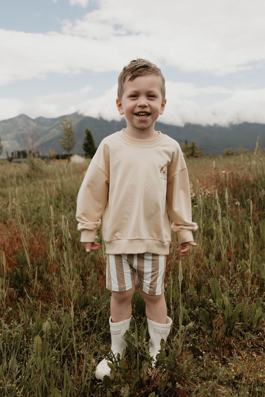 A young child stands in a grassy field with mountains and a cloudy sky in the background. The child is smiling, wearing a beige Children's Sweatshirt by forever French baby made from 95% Cotton and Elastane, striped shorts, and white rain boots, exuding a sense of joy and adventure in the outdoors.