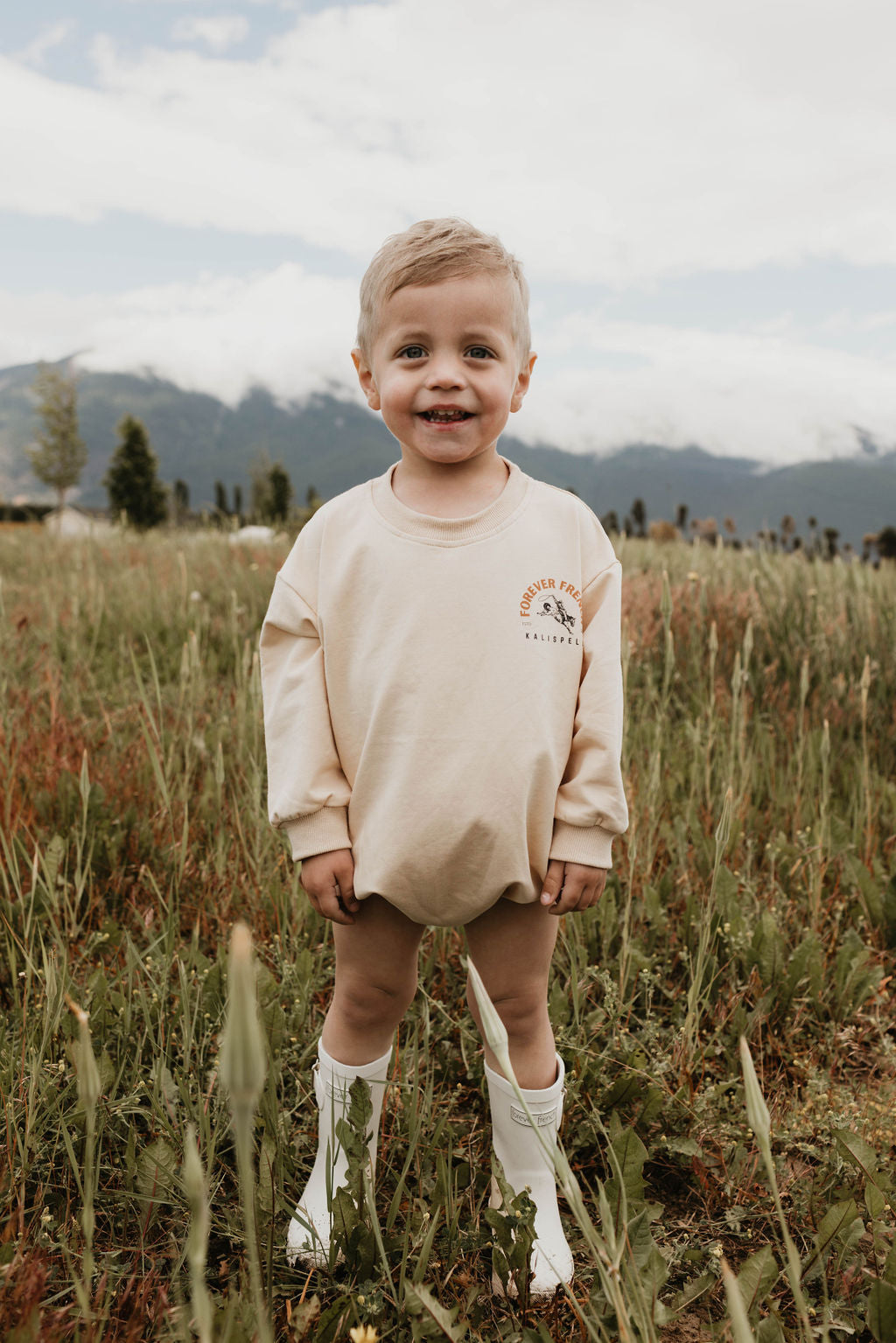 A young smiling child with short light blonde hair stands in a grassy field, wearing the Baby Romper | Kalispell by forever French baby. The child is paired with a diaper and white calf-high boots. The background features mountains under a cloudy sky. Not available for local pickup.