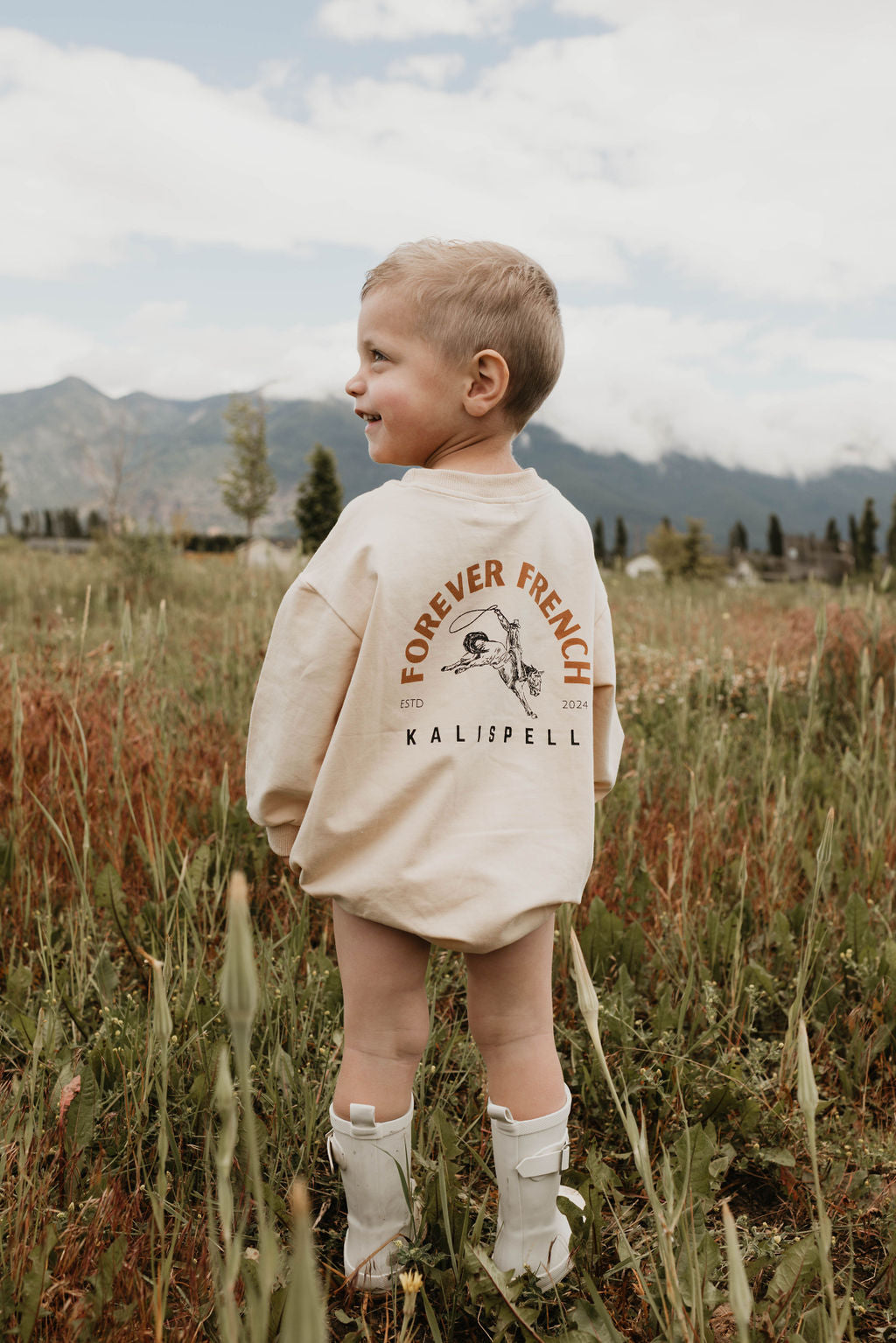 A young child stands in a grassy field with mountains in the background. They are wearing a long beige Baby Romper from forever French baby, made of 95% cotton and elastane, featuring the "Forever French Kalispell" print on the back. The child is also sporting white boots. Looking to the side and smiling under a partly cloudy sky, they seem perfectly content.