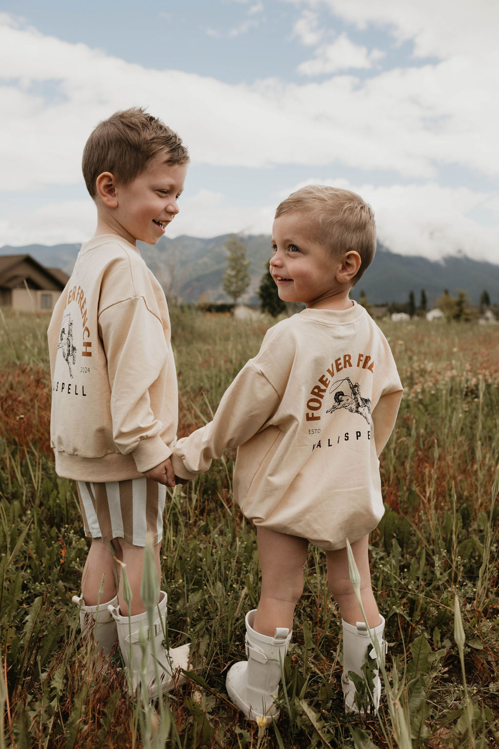 Two young children stand in a grassy field, holding hands and facing each other with smiling expressions. They both wear oversized beige Baby Rompers from the Kalispell collection by forever French baby, made of 95% cotton and elastane; one romper features a logo on the back. They are also wearing white boots. In the background are mountains and a partly cloudy sky.