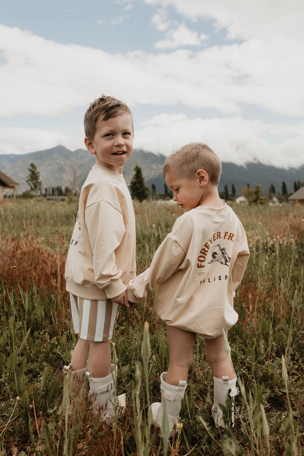 Two young children, both wearing beige Baby Rompers from the forever French baby Kalispell collection made of 95% cotton and elastane, stand hand in hand in a grassy field with mountains in the background. One child is looking back and smiling, while the other gazes down. The rompers have a graphic and text on the back.