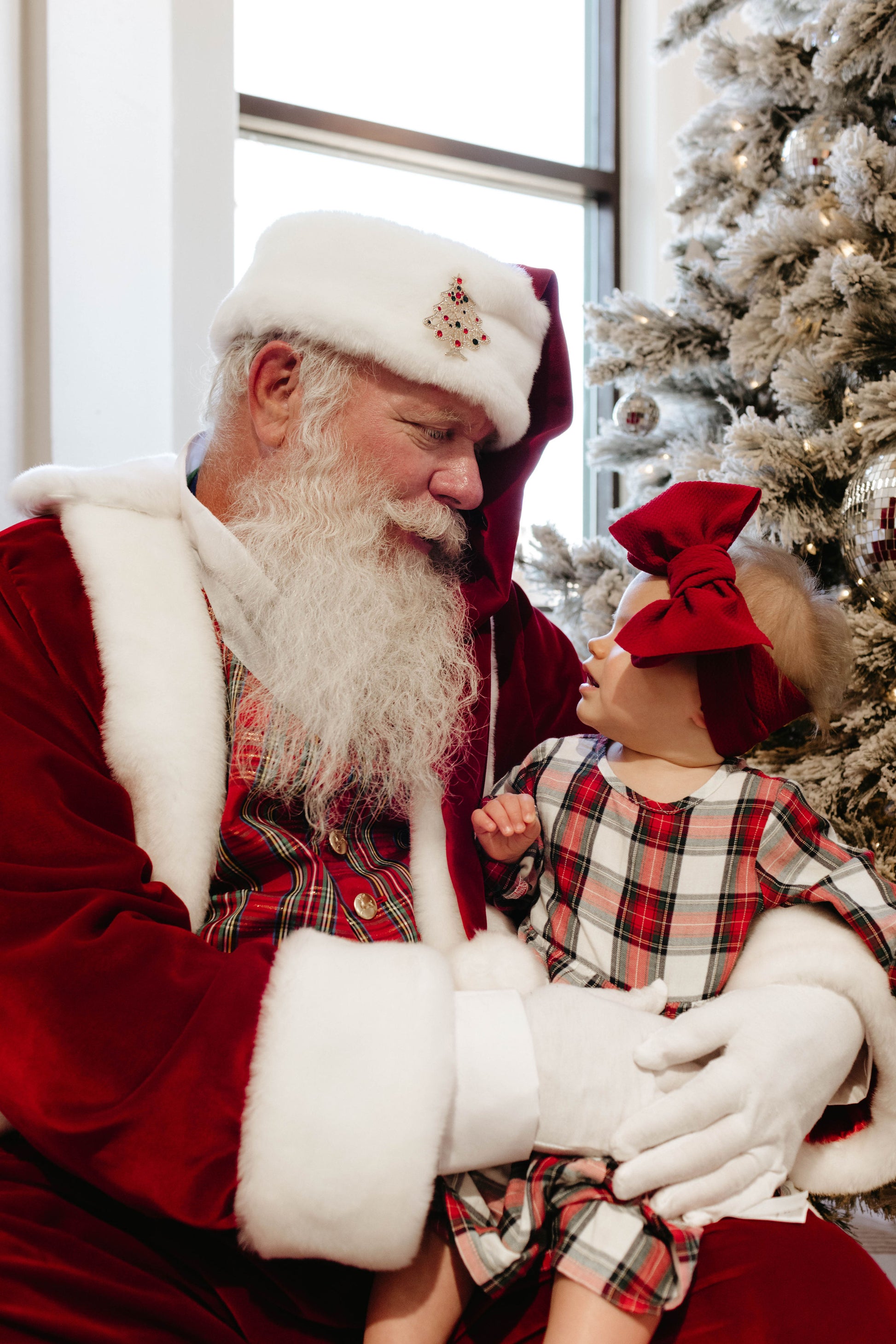 A person dressed as Santa Claus sits next to a decorated Christmas tree, holding a young child in their lap. Capturing this heartwarming moment from the "Santa Photos | Forever French Baby x Amanda Riley Photos" collection, the child wears a red plaid dress and a large red bow on their head. Both Santa and the child are looking at each other and smiling.