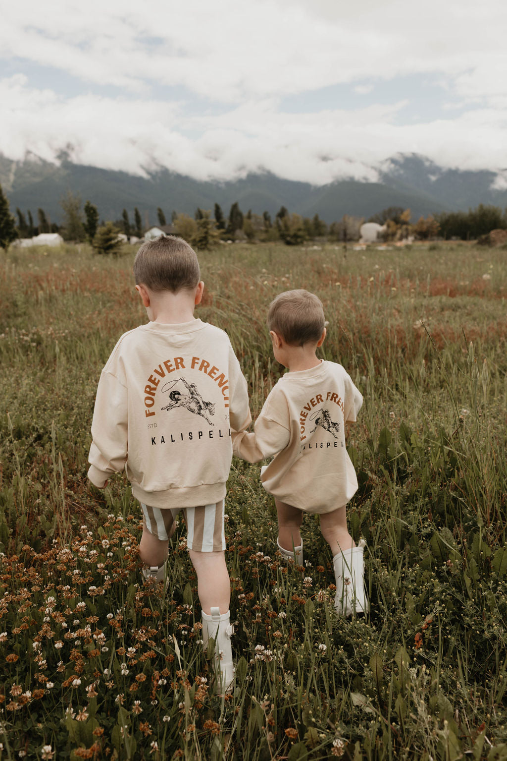 Two young children with short hair walk through a grassy field with mountains in the background. Both children are wearing white boots and cream-colored Baby Rompers from the forever French baby brand, made from 95% cotton, with "Forever French Kalispell" printed on the back. The sky is cloudy, and wildflowers are scattered in the grass.