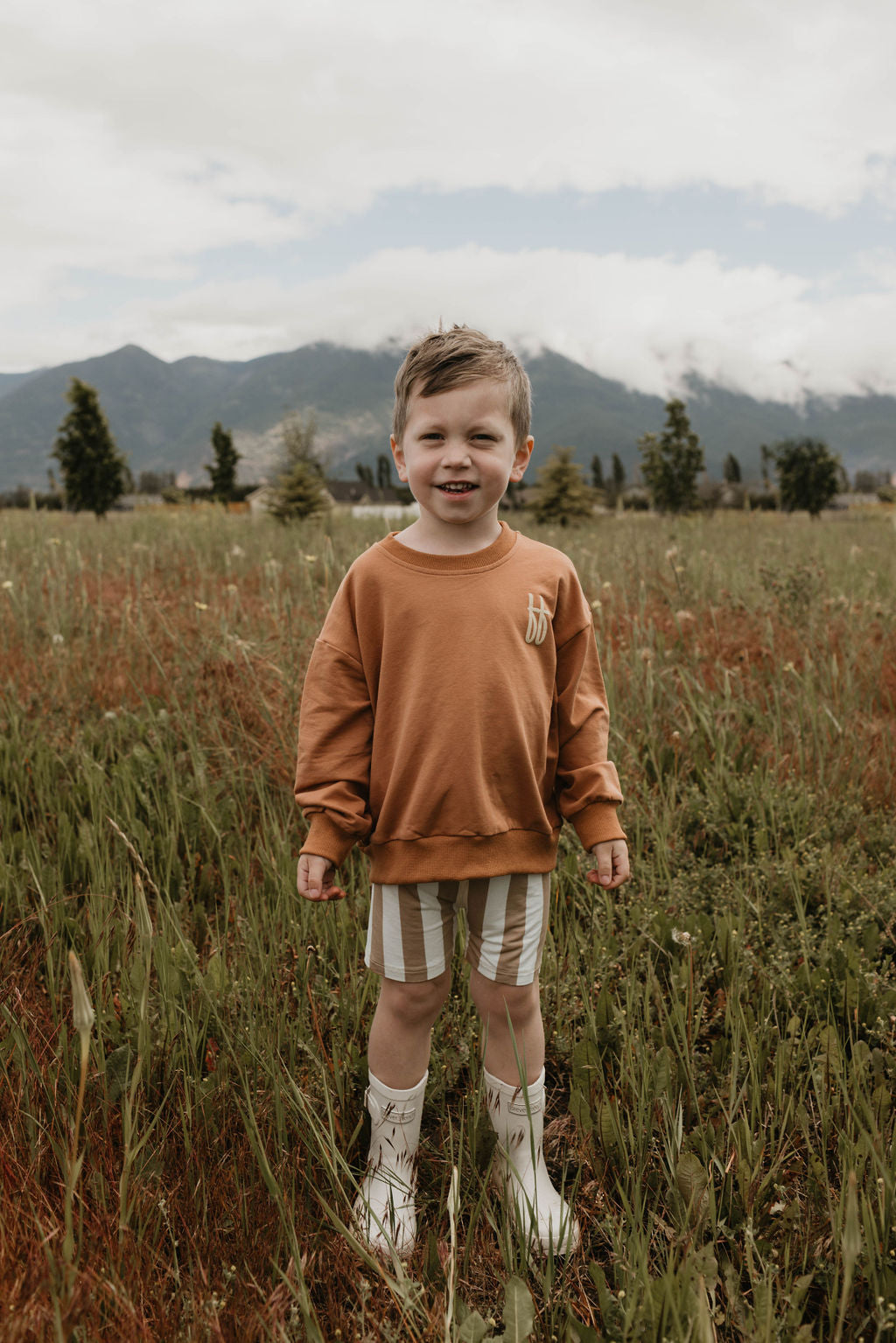 A young boy stands in a grassy field wearing a "Made It to The Mountains" Children's Sweatshirt by forever French baby, made of 95% cotton, paired with striped shorts and white boots featuring a design. He smiles at the camera with mountains and cloudy skies in the background.