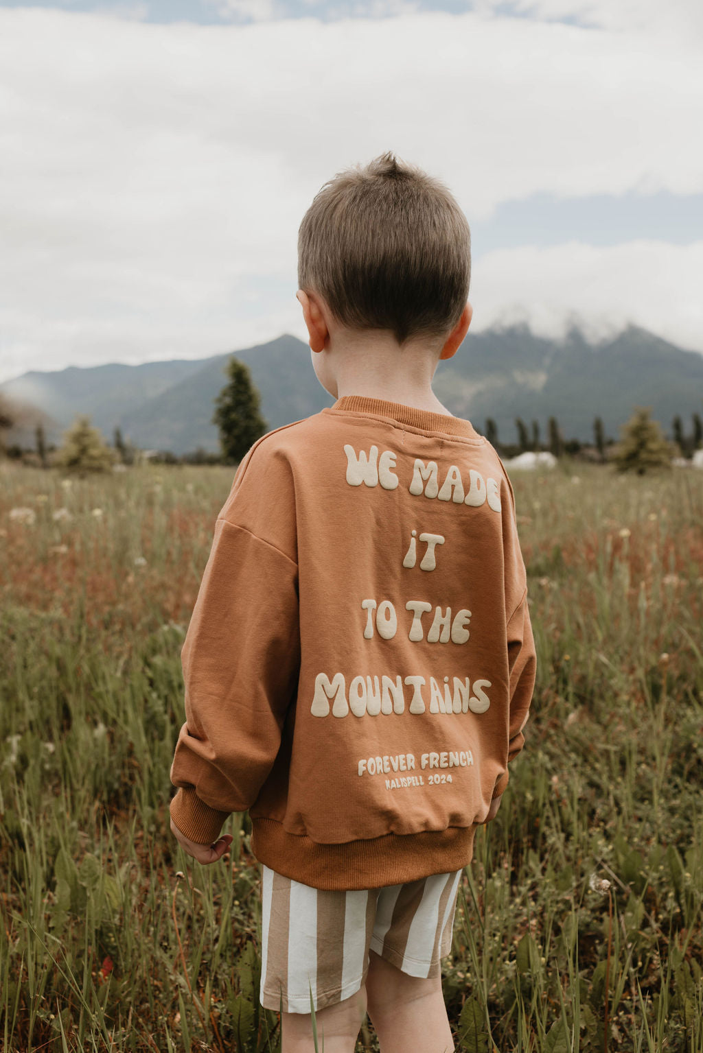 A child with short hair is standing facing away, wearing a brown "Children's Sweatshirt | Made It to The Mountains" from the brand forever French baby. The sweatshirt is made of 95% cotton and elastane and features text that reads, "We made it to the mountains. Forever French Istanbul 2021." The child is in a grassy field with mountains and cloudy skies in the background.