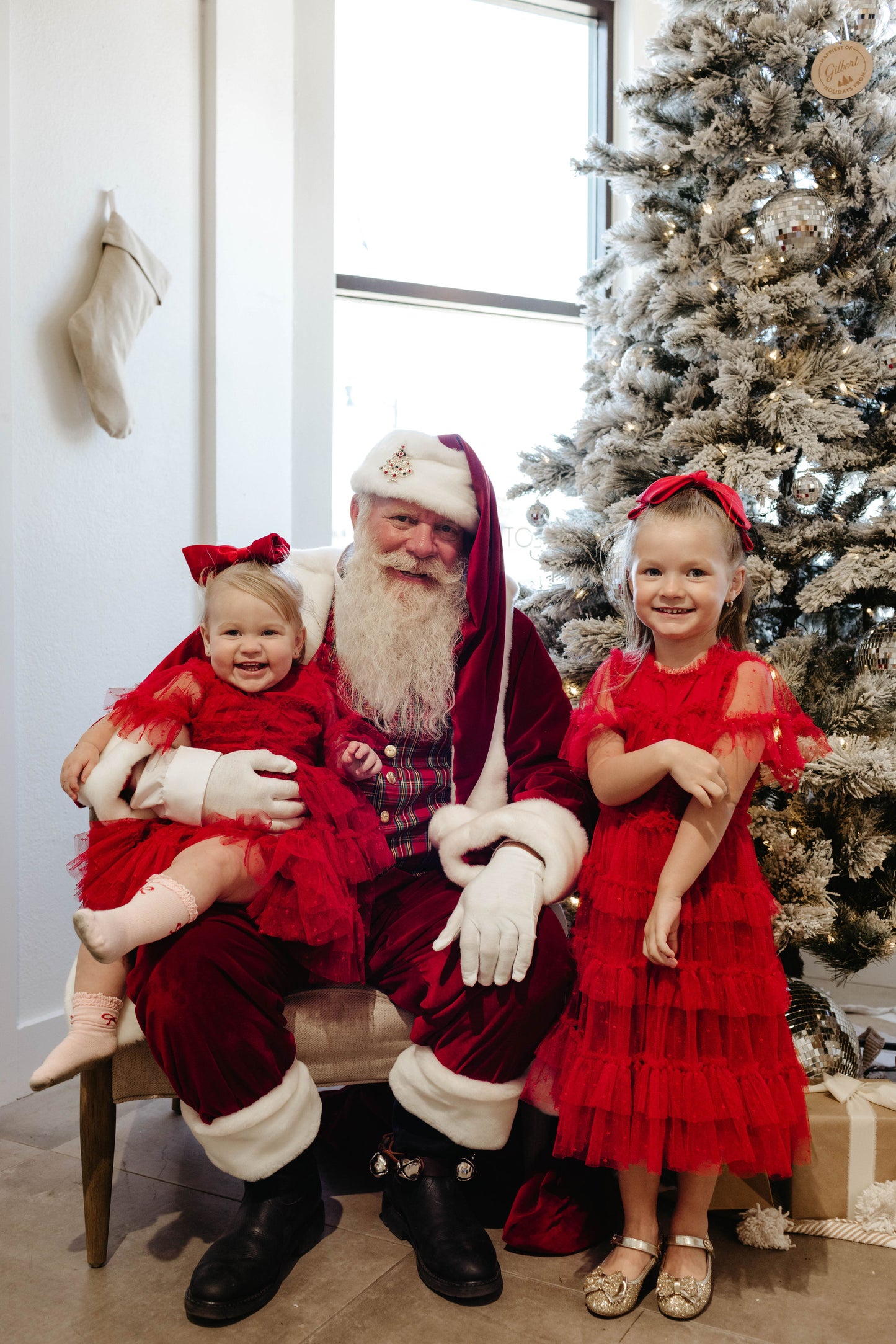 Santa Photos | Forever French Baby x Amanda Riley Photos capture the magic of Christmas as a man dressed as Santa Claus sits with a child in a red dress on his lap and another standing beside him. They pose in front of a white Christmas tree with a stocking hanging on the wall, creating memories to treasure forever.