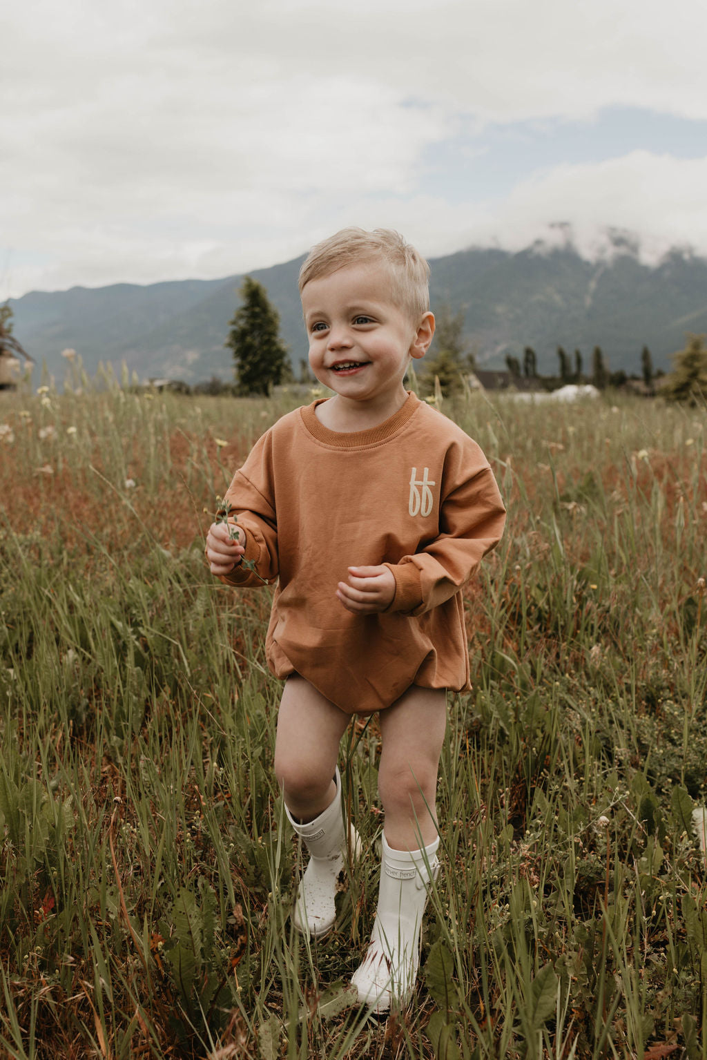 A young child with short blonde hair stands in a grassy field, smiling and holding a small flower. The child is donning an oversized light brown cotton romper from forever French baby, emblazoned with white letters saying "Made It to The Mountains," and is also wearing white boots. The background features mountains partially covered in clouds.