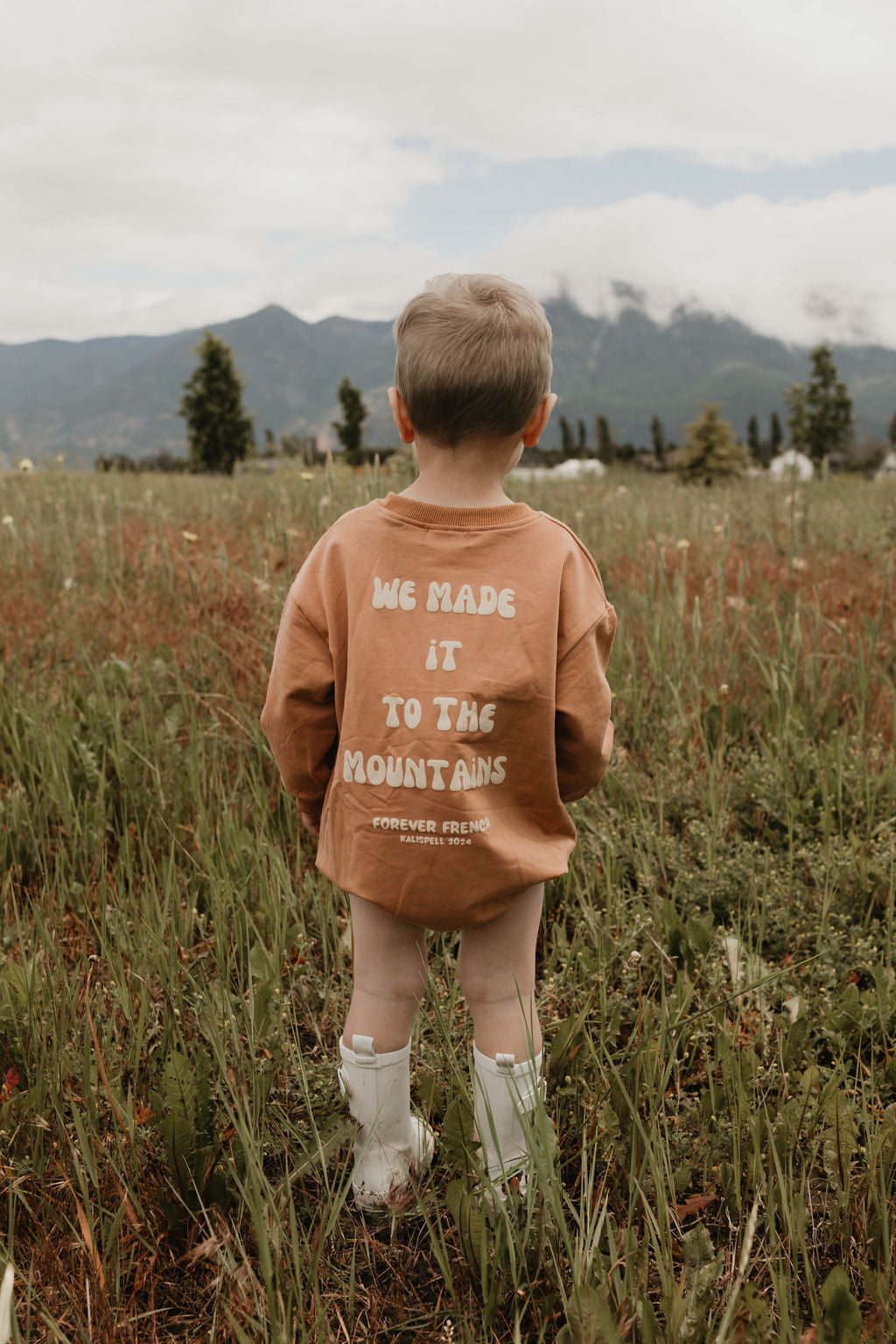 A young child with short hair stands in a field of wildflowers, facing away from the camera. The child is dressed in an oversized tan cotton sweatshirt by forever French baby, featuring the text "We made it to the mountains" on the back and white rain boots. Misty mountains are visible in the background.