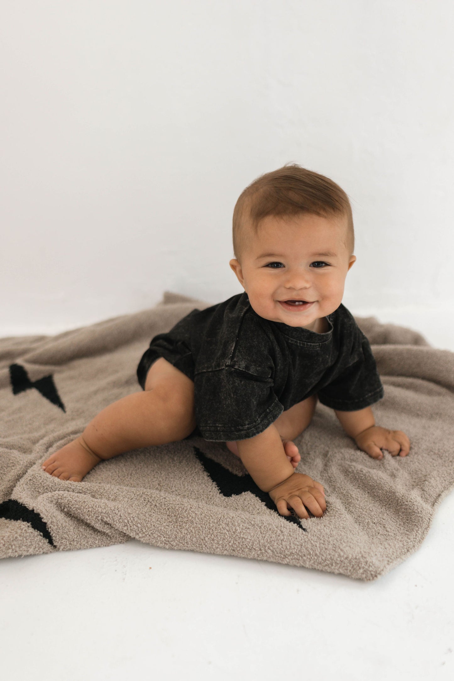 A smiling baby with short, light brown hair wearing a dark, short-sleeved outfit from forever french baby sits on a Plush Blanket in the Brown Lightning Bolt design. The baby is seated on the blanket against a white background with their legs extended and hands resting in front of them.