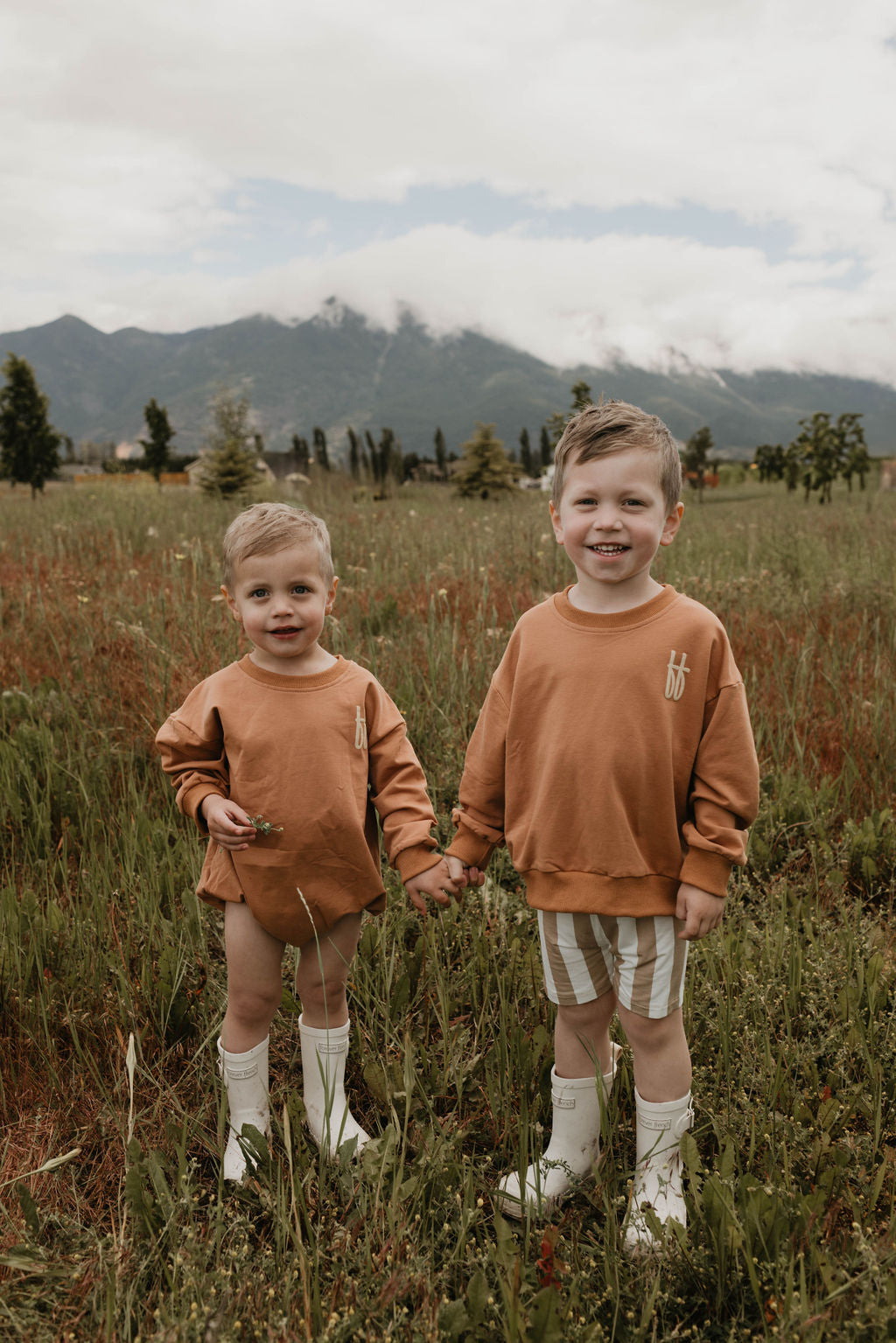 Two young children stand in a grassy field with mountains in the background. Both wear matching Baby Romper | Made It to The Mountains sweatshirts from forever French baby, crafted from soft cotton fabric, paired with white boots. One child has shorts, while the other wears striped shorts. They are holding hands and smiling at the camera.