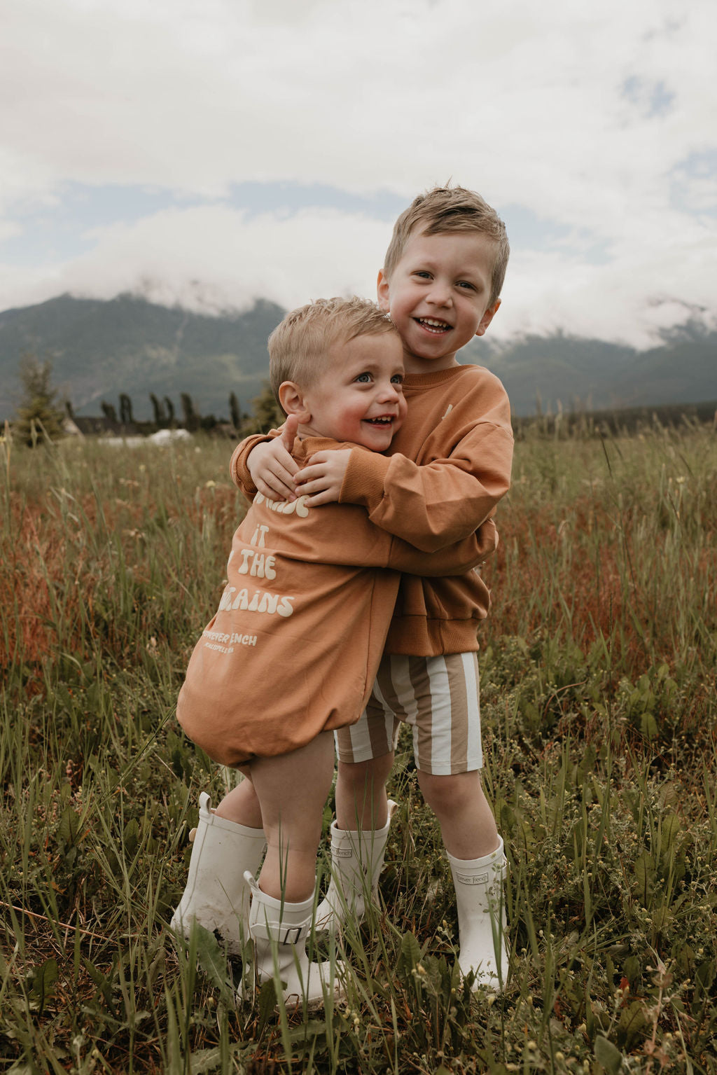 Two young boys are standing in a grassy field, hugging each other and smiling at the camera. They are both wearing the "Made It to The Mountains" sweatshirts by forever French baby, made from 95% cotton and elastane, paired with white rain boots. The background features mountains and a cloudy sky.