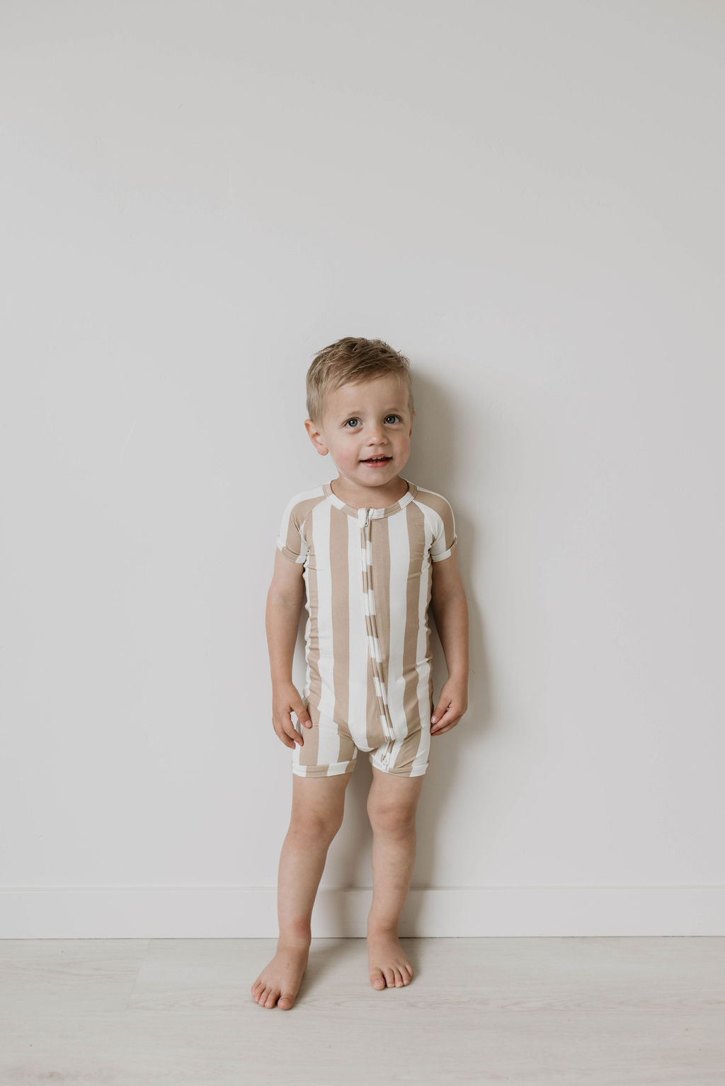 A young child with blonde hair stands barefoot against a plain white wall, wearing the "Shortie Bamboo Zip | Kal Stripe" romper by forever french baby. The hypo-allergenic fabric features short sleeves and beige and white stripes. There's a slight smile on their face.