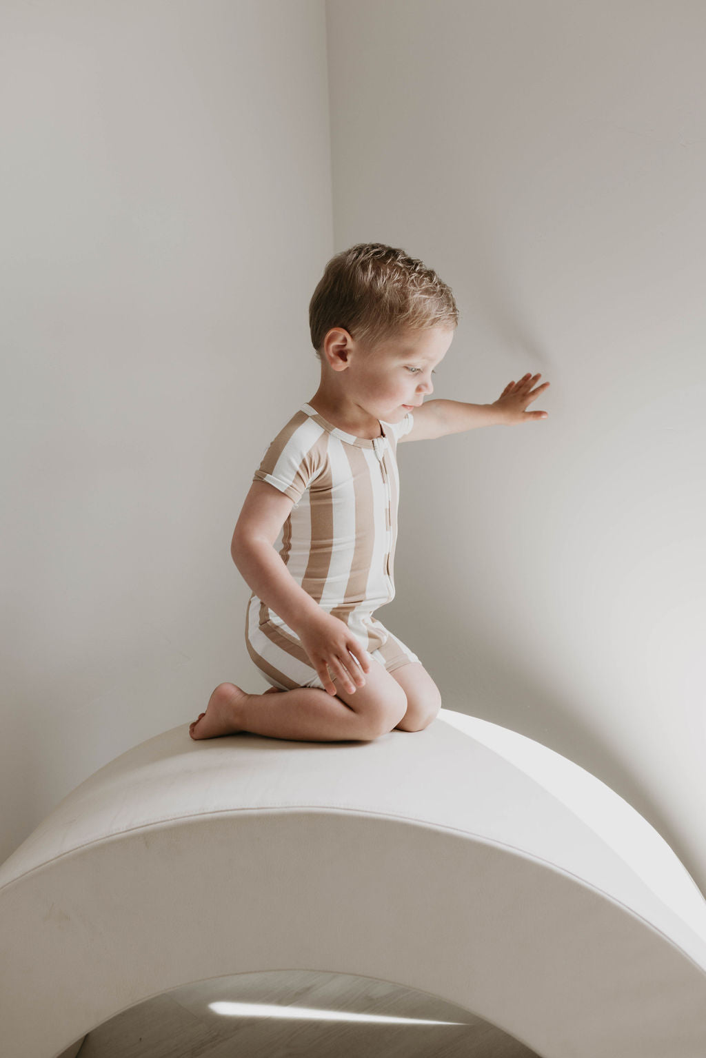 A young child with short hair, dressed in the "Shortie Bamboo Zip | Kal Stripe" baby romper from forever french baby, kneels on a curved, white surface. The child reaches out with one hand towards a sunlit wall, appearing curious and engaged. The setting is minimalist with soft natural light.