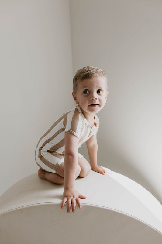 A young child with short blonde hair kneels on a white surface, wearing the Shortie Bamboo Zip in Kal Stripe by Forever French Baby. The child gazes upward with a curious expression while soft, natural light illuminates the scene against a plain, light-colored background.