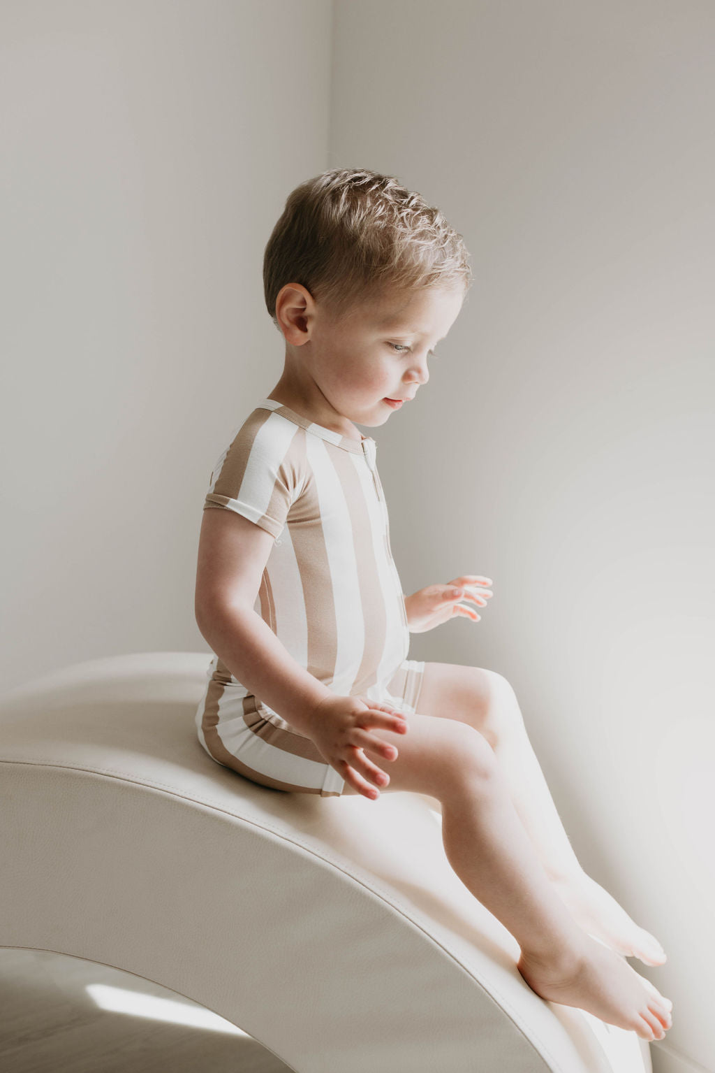 A young child with short, light hair is sitting on a curved, light-colored surface. They are dressed in a **Shortie Bamboo Zip | Kal Stripe** baby romper from **forever french baby**, featuring soft beige and white stripes. Soft natural light bathes the scene, enhancing its calm and serene atmosphere. The child looks downward, appearing thoughtful.