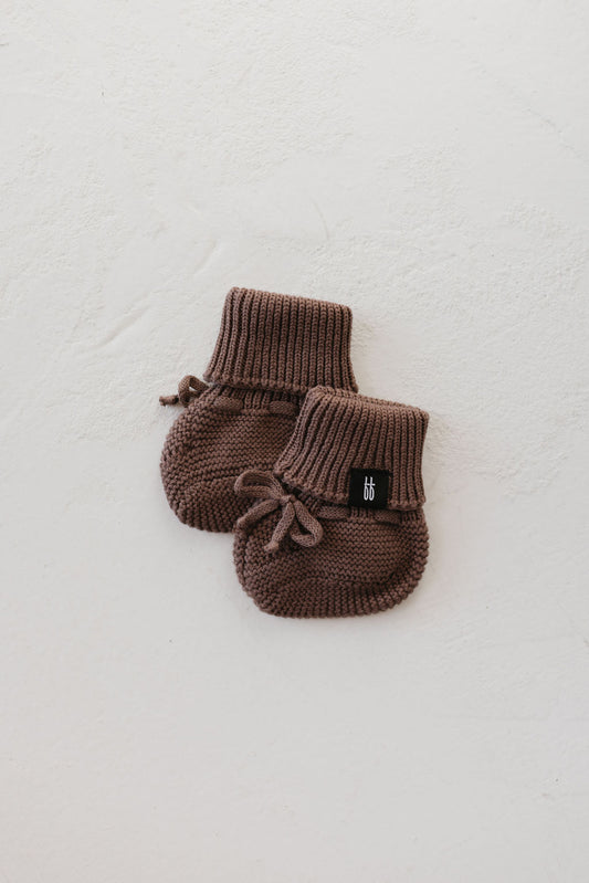 A pair of ff Knit Booties in the Chocolate Chip color from forever french baby, featuring ribbed cuffs and ties, laid on a textured white surface. A small black label is attached to the booties.