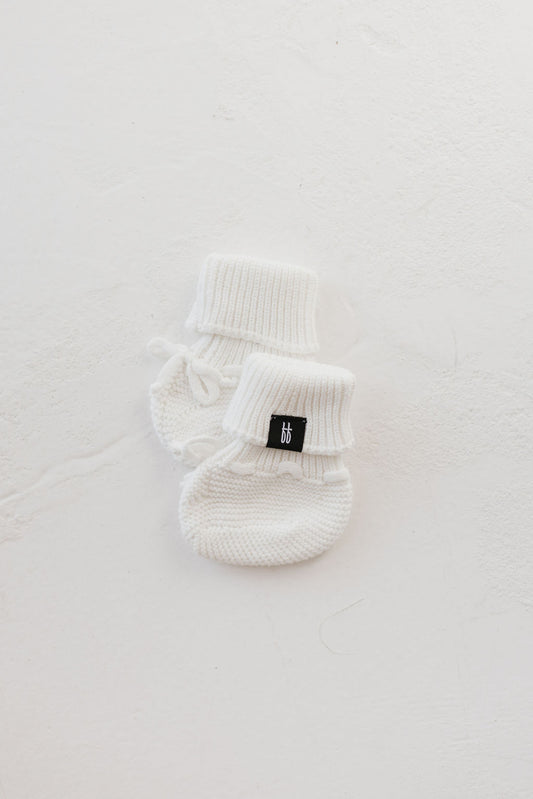 A pair of ff Knit Booties in Snow by forever french baby, featuring folded cuffs, resting on a smooth white surface. A small black tag is attached to one bootie.