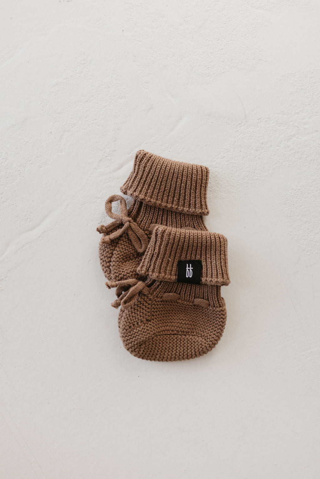 A delightful pair of caramel chip knit booties from forever french baby, complete with ribbed cuffs and small ties, sits elegantly on a textured white surface—ideal for the cozy winter months.