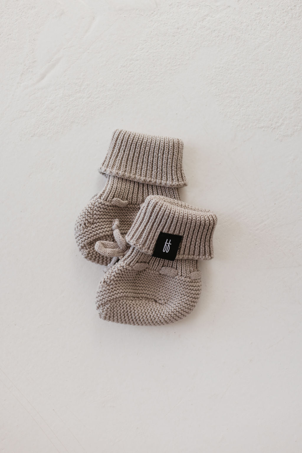 The ff Knit Booties in Silver Bell from forever french baby feature snug fold-over cuffs, making them ideal for the winter months. One bootie is decorated with a small black tag, beautifully contrasted against a light textured background.