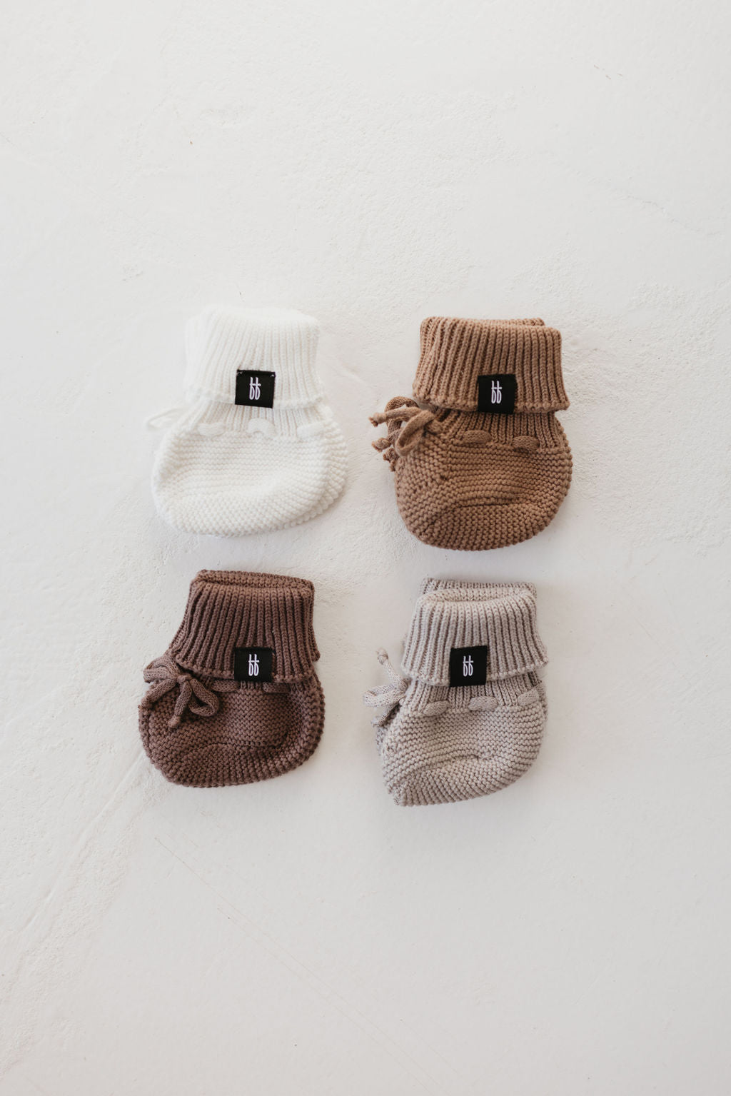 Four pairs of ff Knit Booties by forever french baby are arranged in a square on a light surface. The booties come in white, tan, brown, and light brown shades, each featuring a small black label and ribbed cuffs.