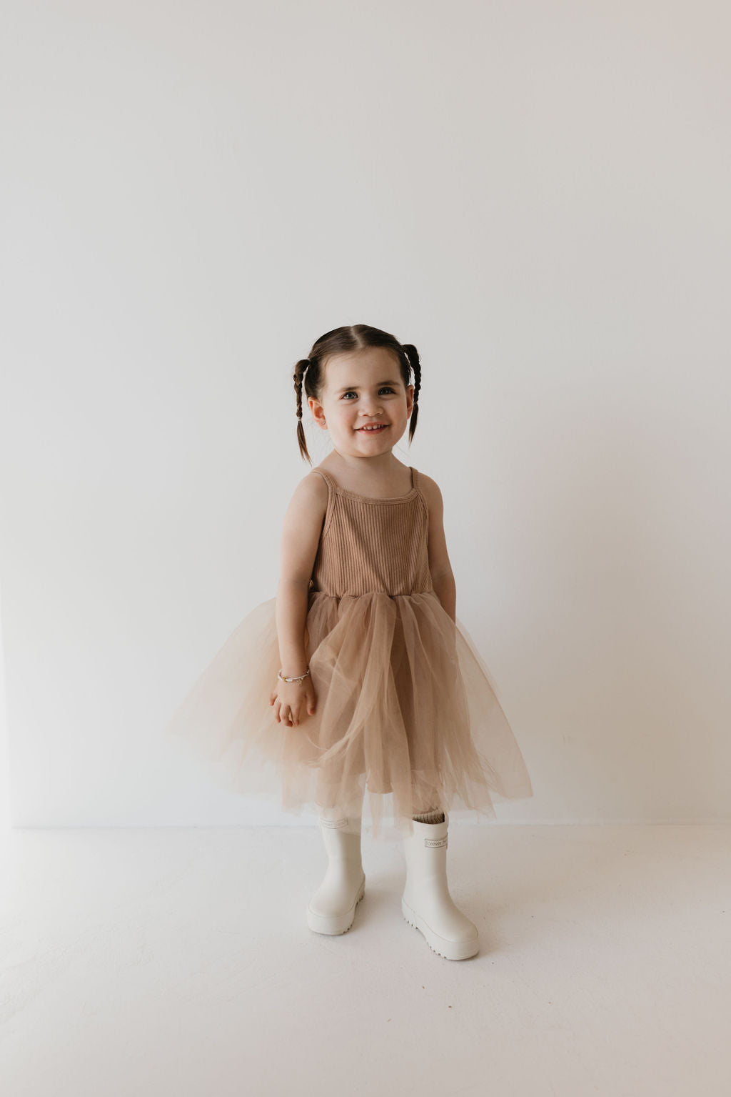 A young girl with pigtails beams in her Spice ff Tutu Dress by forever french baby, paired with charming white boots. She stands against a plain white backdrop.