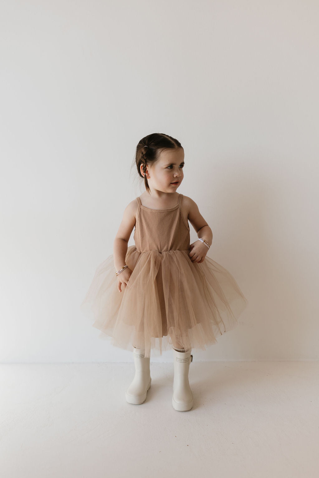 A young child stands against a plain background, wearing the ff Tutu Dress in Spice from forever french baby, paired with white rain boots. The child has short, dark hair.