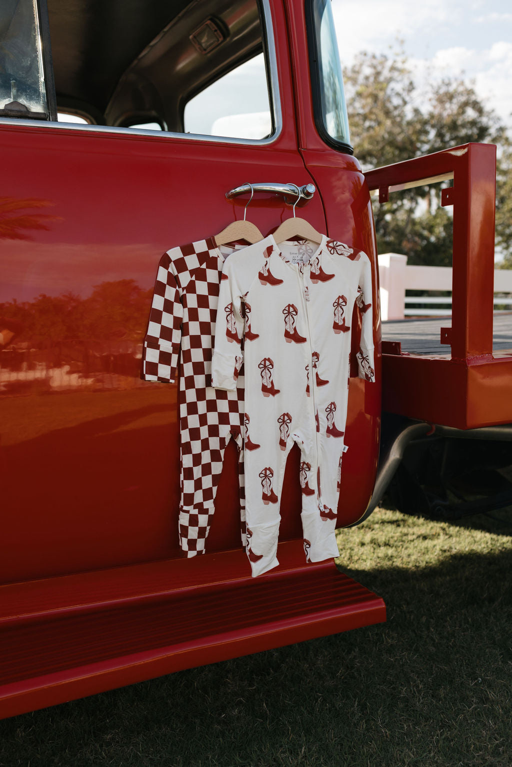 Two onesies from lolo webb's Bamboo Zip Pajamas collection hang on wooden hangers from a red vintage truck, highlighting their hypo-allergenic fabric. One showcases a red and white checkerboard design, while the other displays playful red dogs against a white backdrop. Set outdoors, the trees and blue sky provide the perfect setting for these Cowgirl Christmas garments.