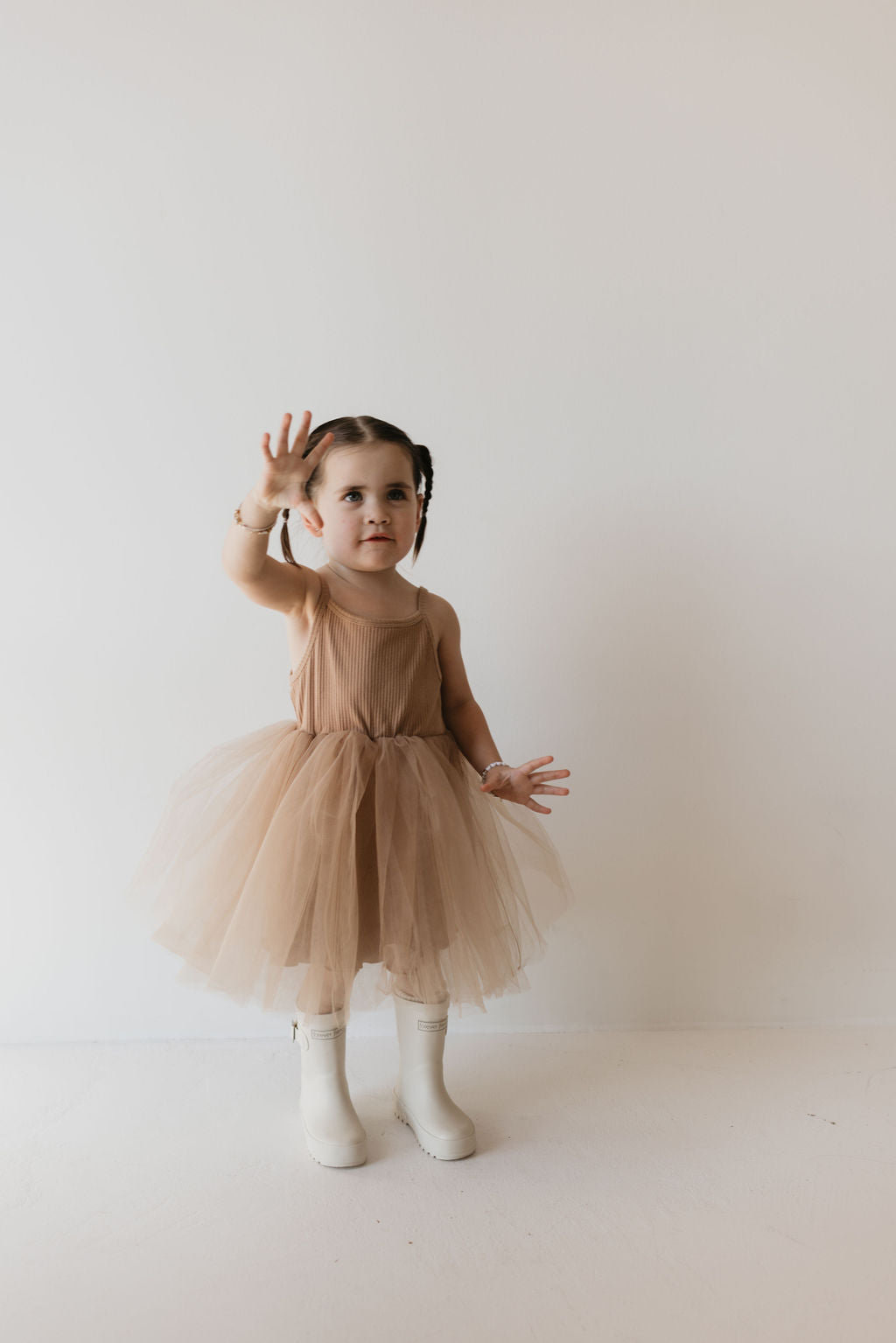 A young girl with braided hair is wearing the ff Tutu Dress in Spice by forever french baby and white rain boots. She stands against a plain light background, with one arm raised slightly, as if reaching or waving.