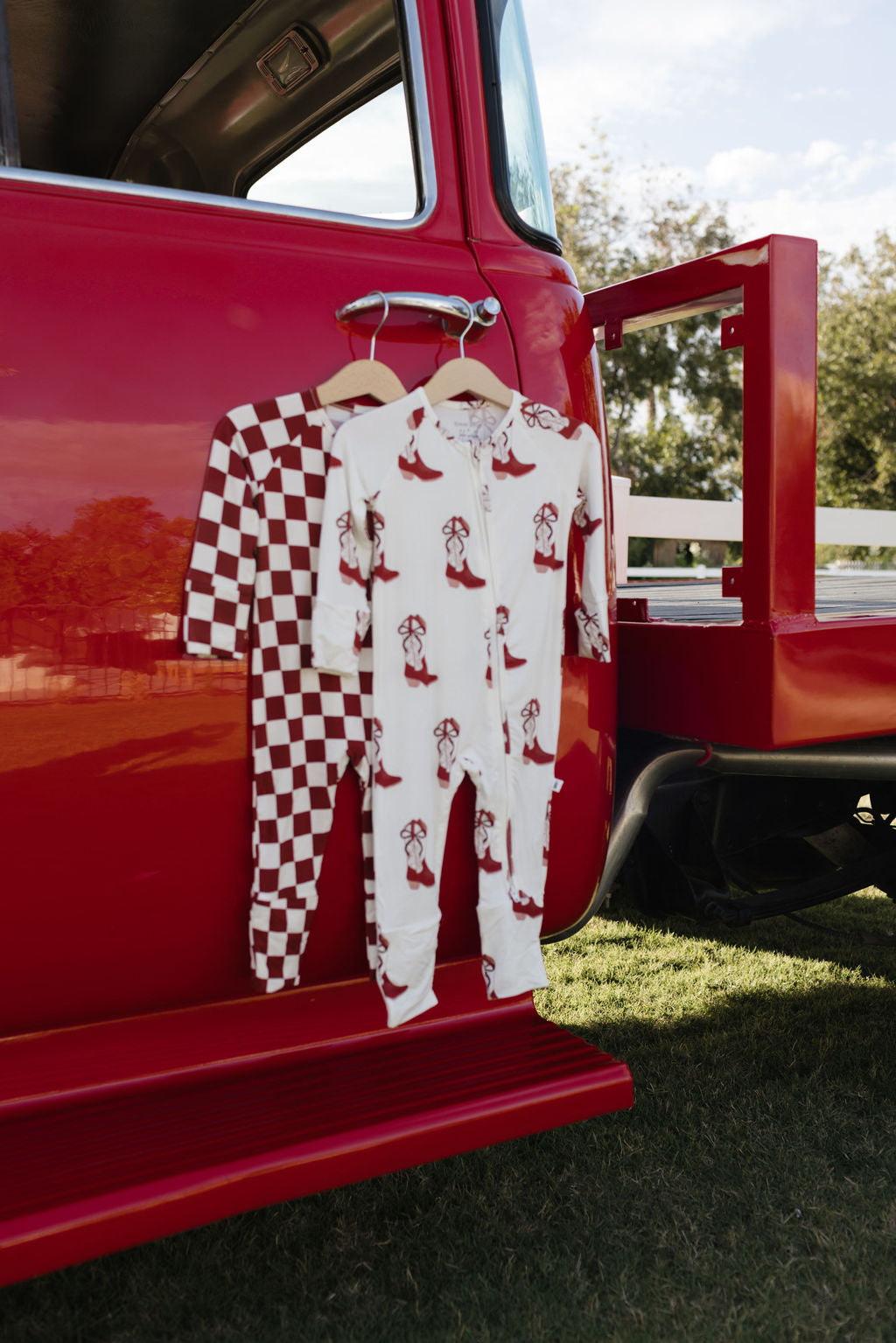 Two stylish onesies from lolo webb hang on a red truck. The Bamboo Zip Pajamas in Cowgirl Christmas design feature a hypo-allergenic, red and white checkered pattern, while the other piece with penguin patterns serves as breathable sleepwear. The scene unfolds outdoors with grass and trees in the backdrop.