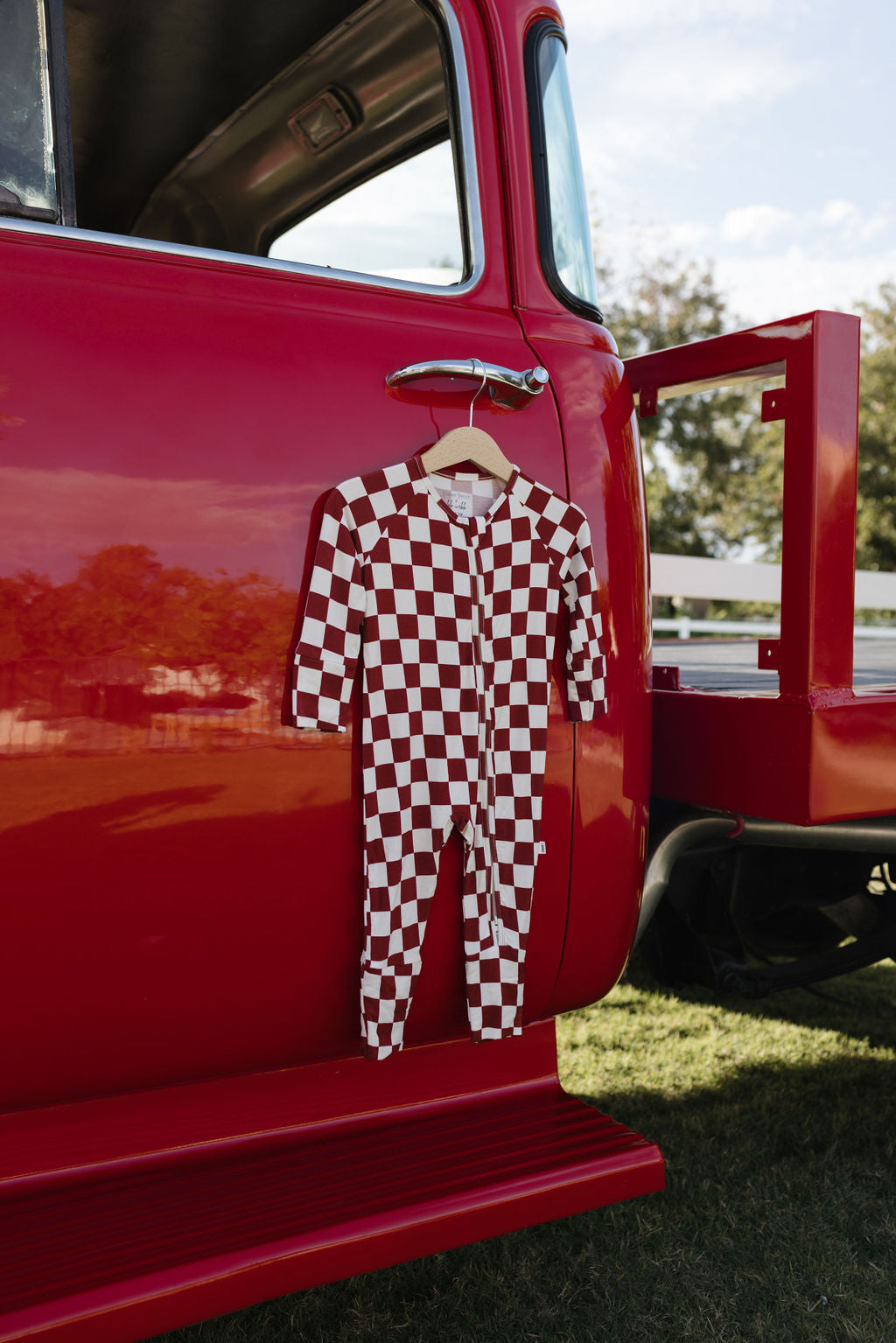 A red and white checkered hypo-allergenic baby jumpsuit, known as the Bamboo Zip Pajamas | the Quinn by lolo webb, hangs on a hanger from the open door of a vintage red truck. The truck is parked on grass, with a bright sky and trees in the background.