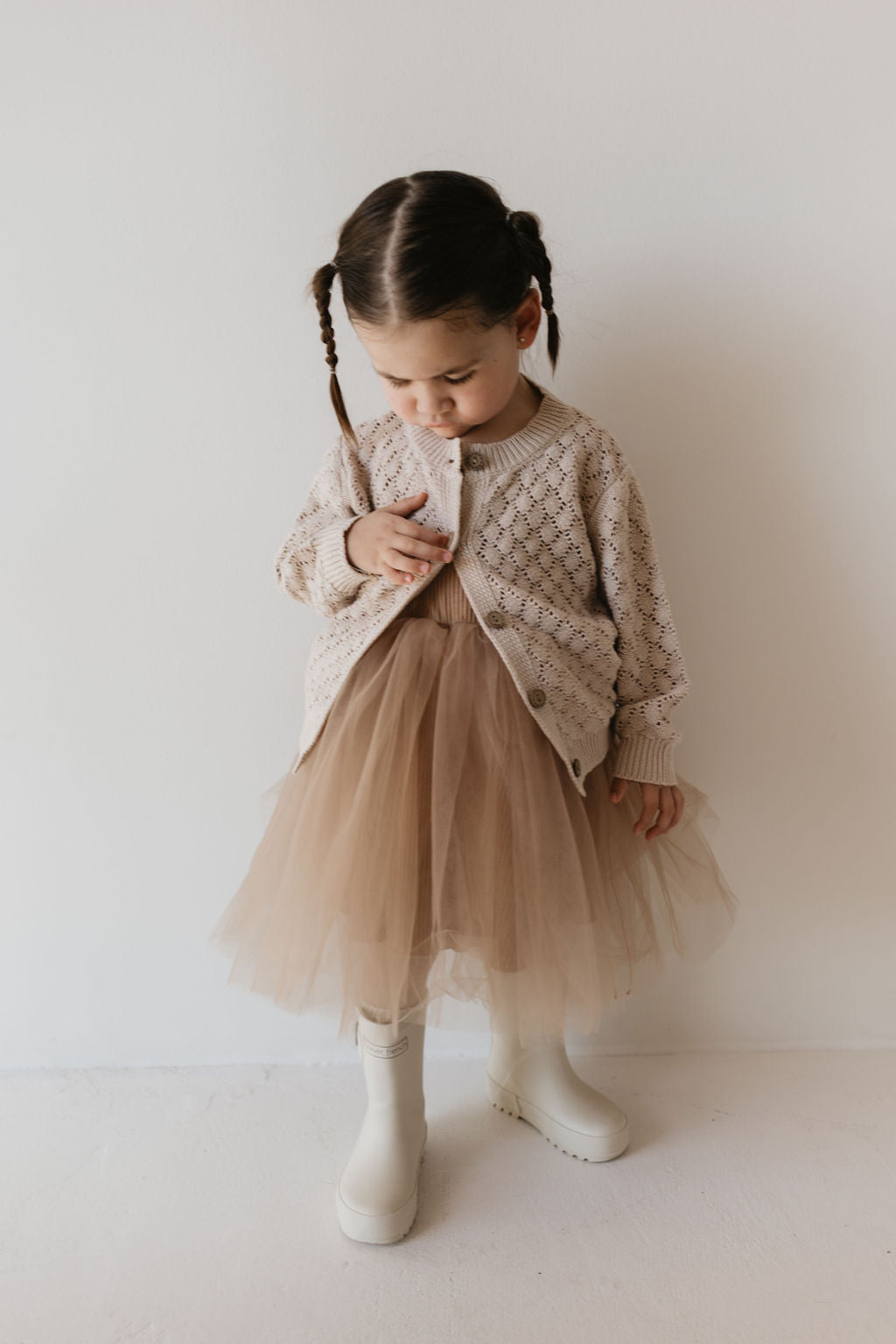 A young child stands against a plain background, looking down. They wear a warm-toned Knit Cardigan in Biscuit by forever french baby, paired with a light brown tutu skirt and white boots. Their hair is styled in two braided pigtails.