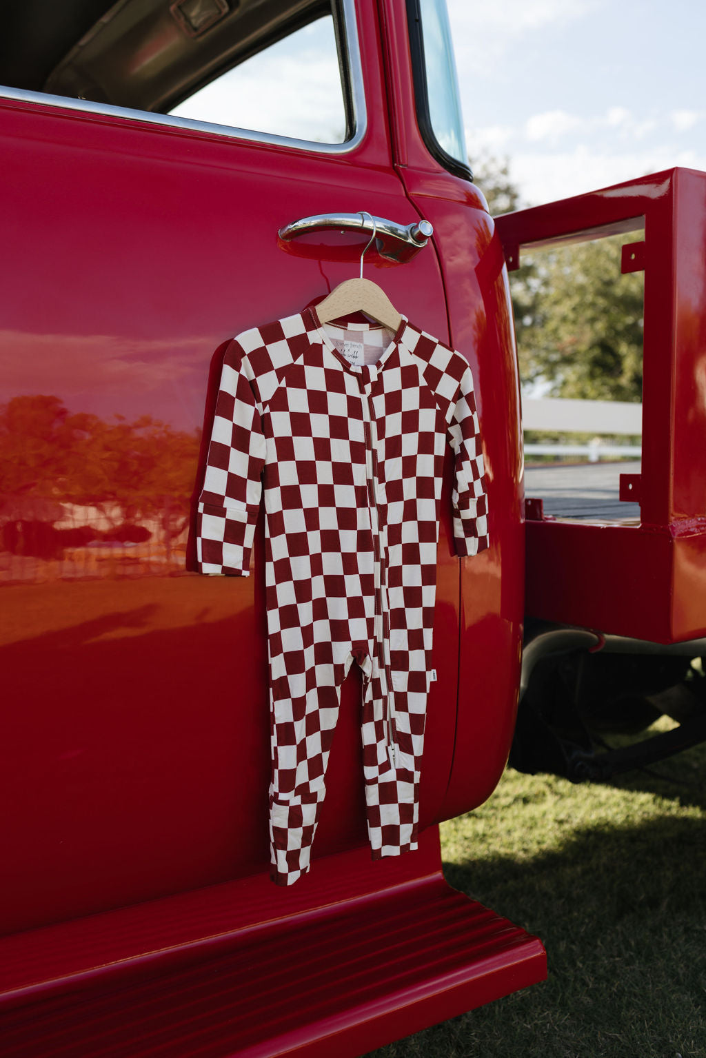 A onesie from lolo webb, the Bamboo Zip Pajamas | the Quinn, adorned in a red and white checkered design and made from hypo-allergenic material, hangs on a hanger from the open door of a vintage red truck parked on grass. Sunlight dances across it as trees cast shadows in the background.