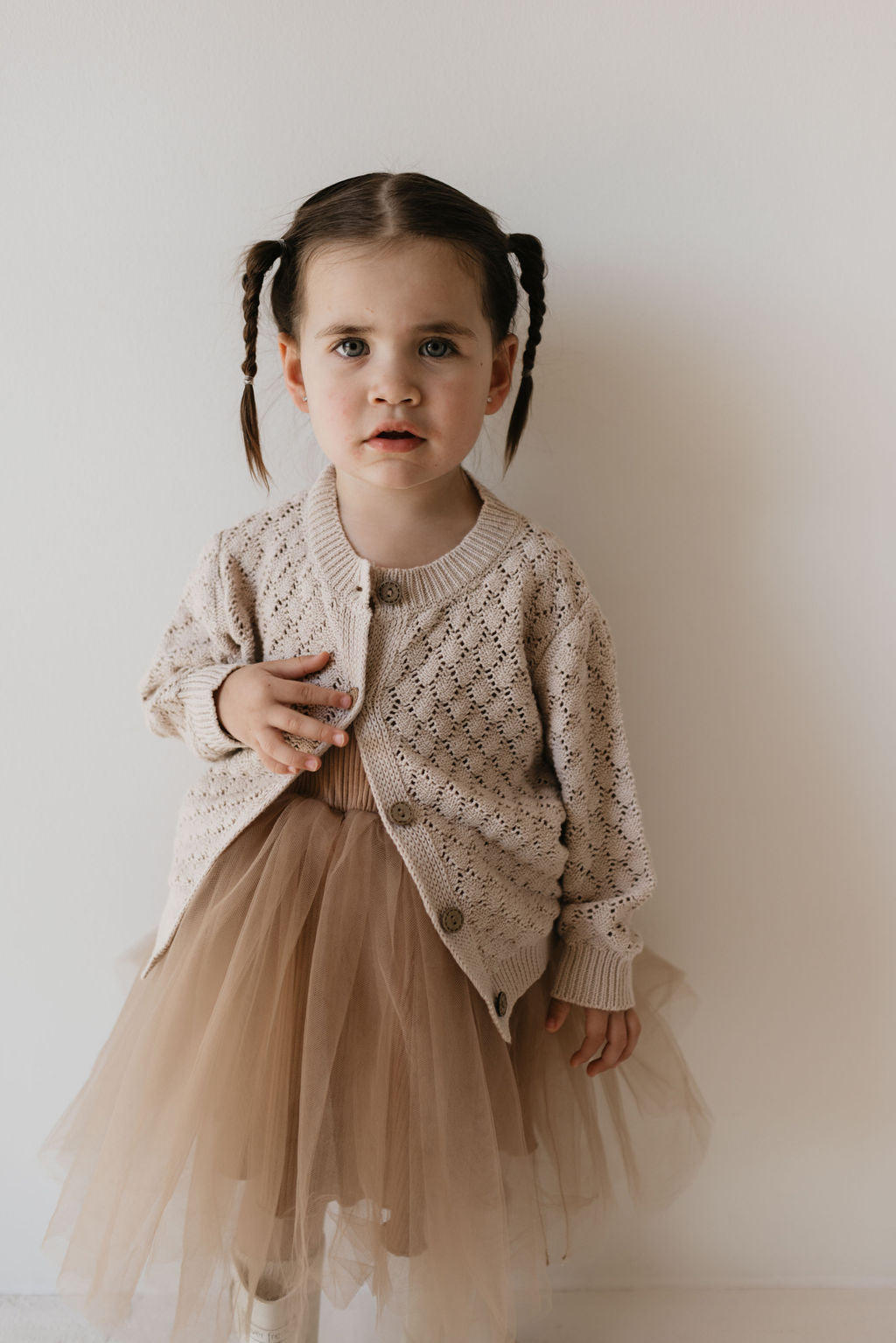 A young girl with braided hair is wearing a knit cardigan in the shade Biscuit by Forever French Baby and a brown tulle skirt. She stands against a plain wall, looking towards the camera.