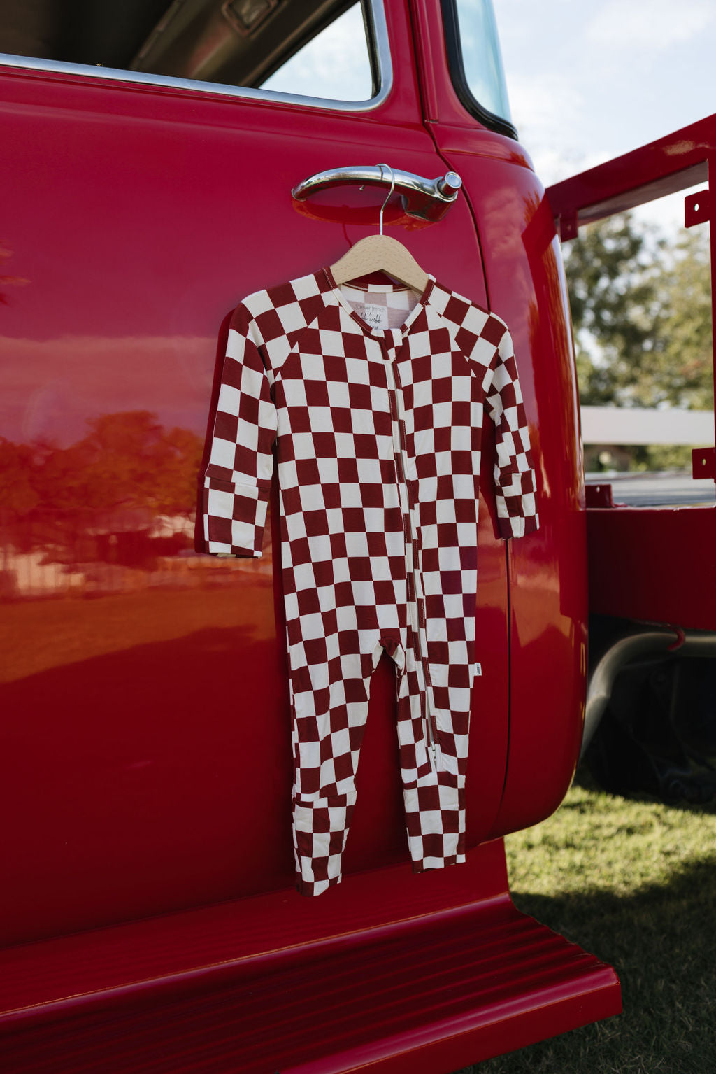 A Bamboo Zip Pajama in the Quinn design by lolo webb, featuring a red and white checkered pattern made from breathable baby wear fabric, hangs on the red door handle of a vintage vehicle. The scene is set outdoors, with grassy areas and trees visible in the background.