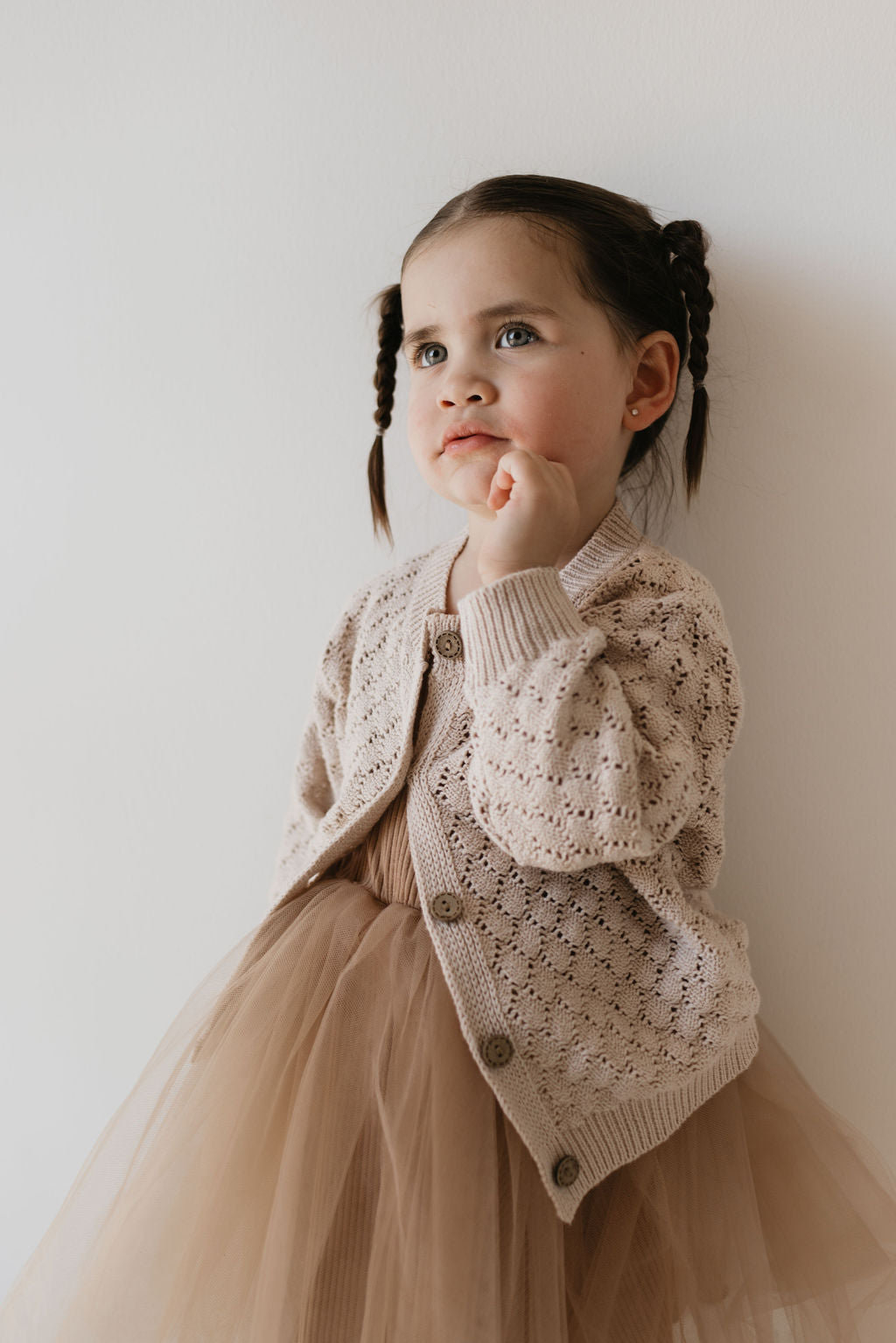 A young child with dark hair in pigtails wears the "Knit Cardigan | Biscuit" from Forever French Baby over a light brown tulle dress. The child stands against a plain wall, looking thoughtfully to the side, with a finger resting on their chin.