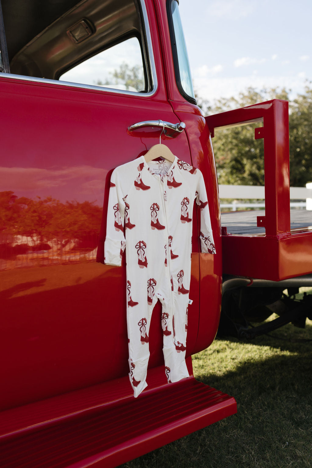 A red pickup truck with the Bamboo Zip Pajamas | Cowgirl Christmas, a hypo-allergenic onesie from lolo webb featuring red dog patterns, hangs on a wooden hanger from the open door. The truck is parked on grass, framed by a green tree and a white fence in the background.