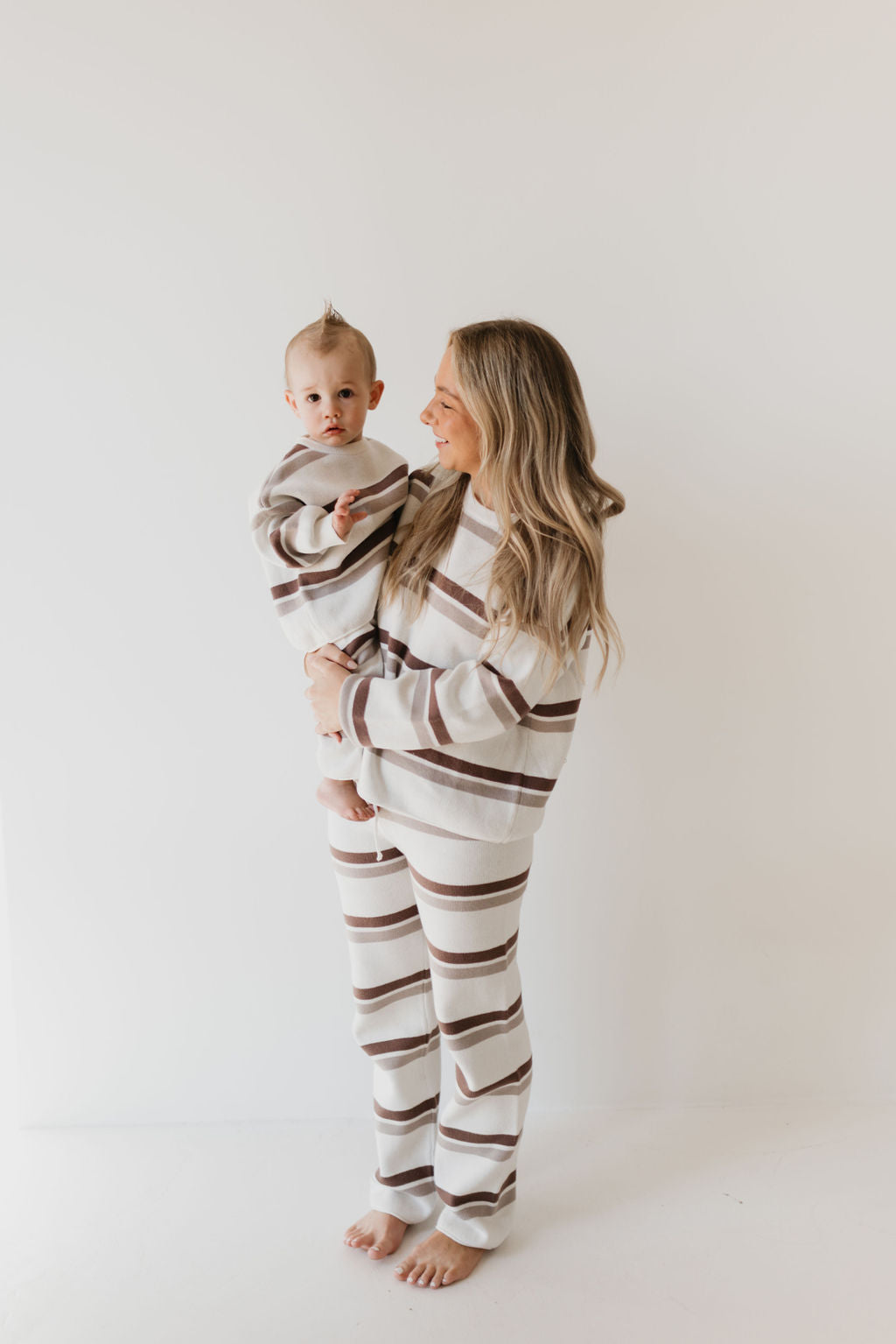 A woman and a child are both dressed in matching Woodland Stripe Knit Pant Sets from forever french baby, making a perfect addition to their fall wardrobe. The 100% cotton fabric ensures comfort as the woman holds the smiling child against a plain white background.