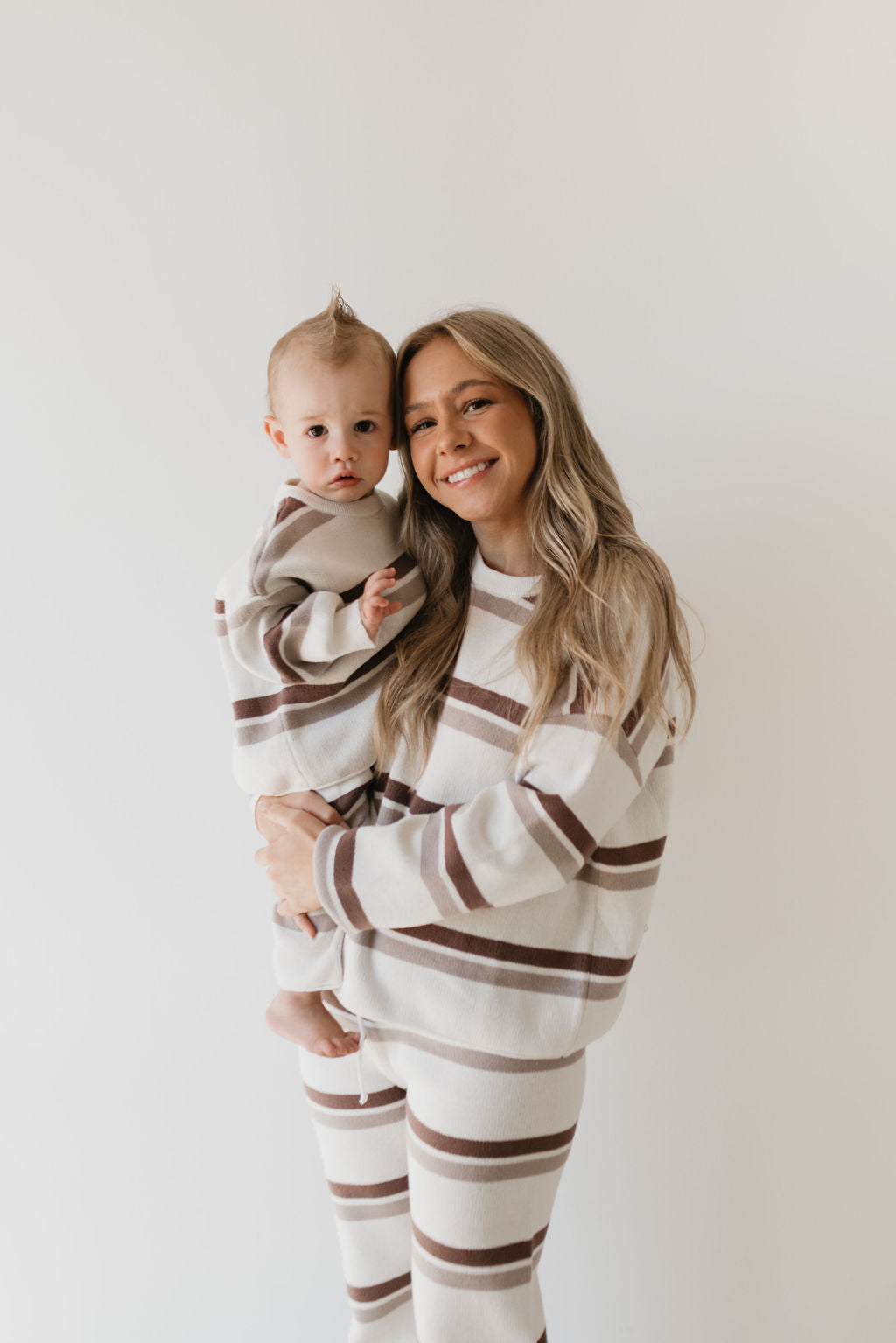 A woman and her baby, both beaming in their matching Knit Pant Set | Woodland Stripe from forever french baby, are dressed in beige and brown striped outfits. The woman with long blonde hair holds the baby sporting an adorable mohawk. Their cozy attire is ideal for a fall wardrobe against the minimalist white background.