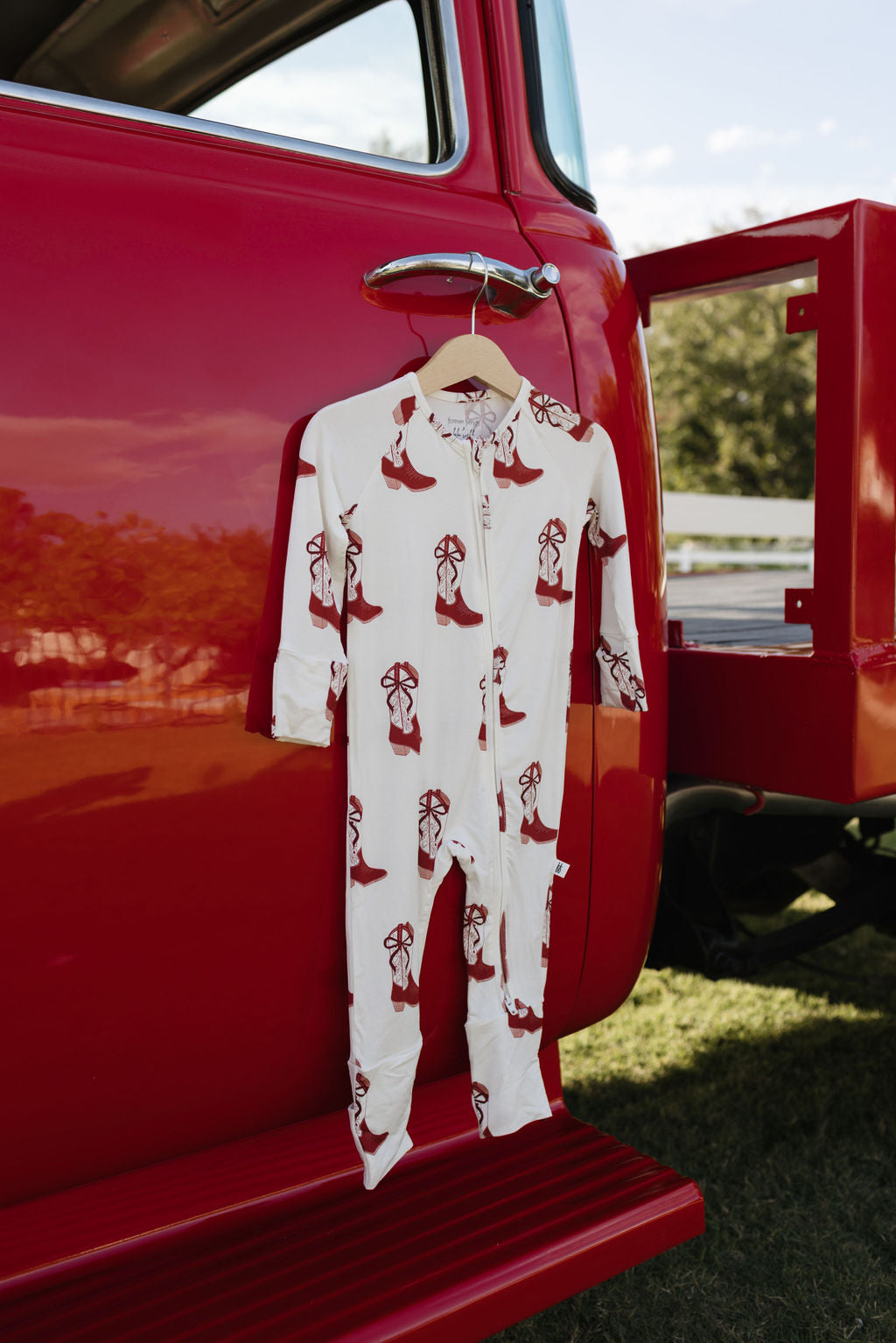 A pair of bamboo zip pajamas from lolo webb, featuring a festive Cowgirl Christmas design, drapes over the door handle of a red vintage truck parked outside. Made from breathable, hypo-allergenic material, it enjoys the clear sky's embrace with a white fence nearby.