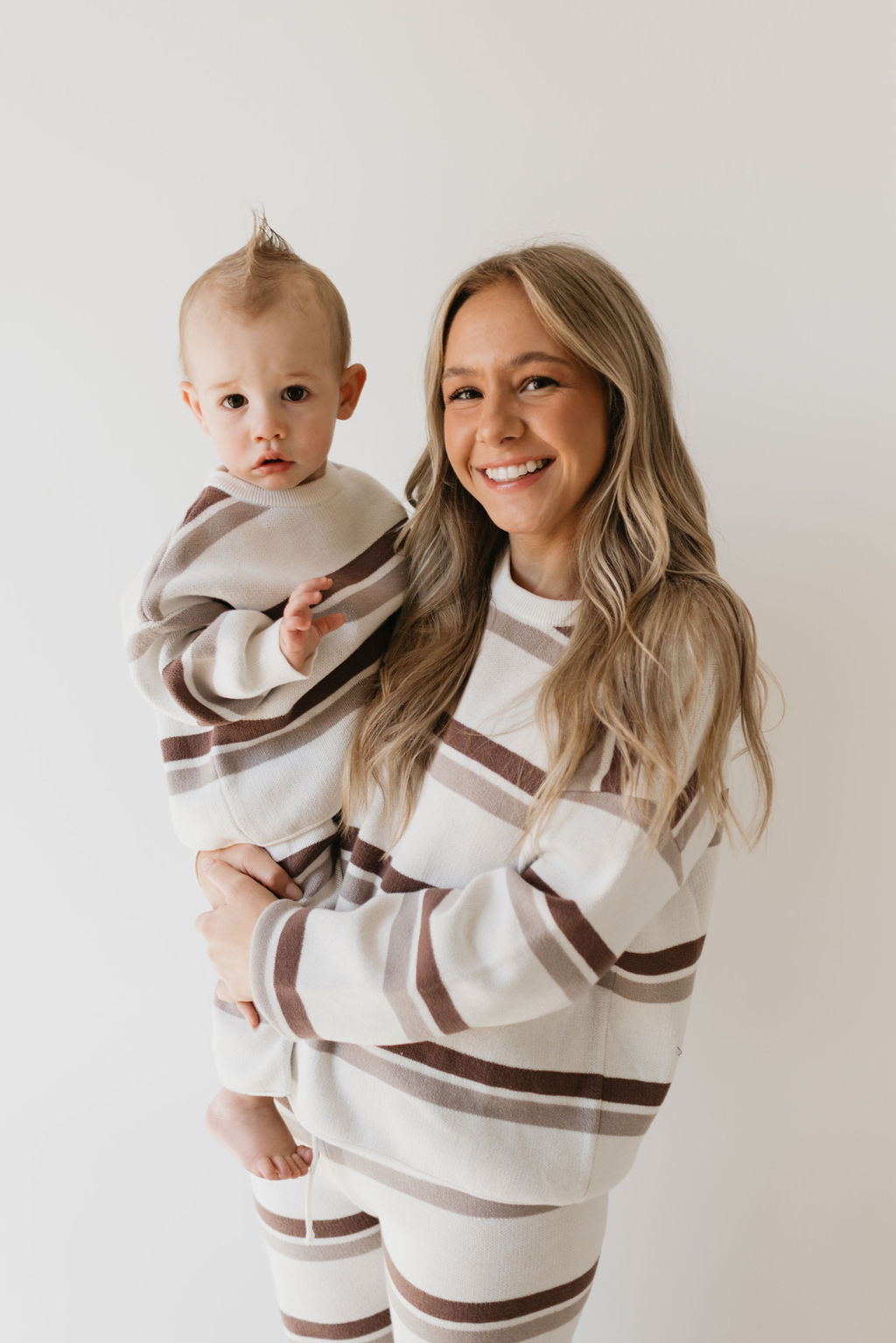 A woman with long blonde hair holds a child, both dressed in the Knit Pant Set | Woodland Stripe from forever french baby, showcasing matching 100% cotton outfits with brown and white horizontal stripes. Their cozy attire perfectly complements the plain white wall behind them as they warmly smile at the camera.